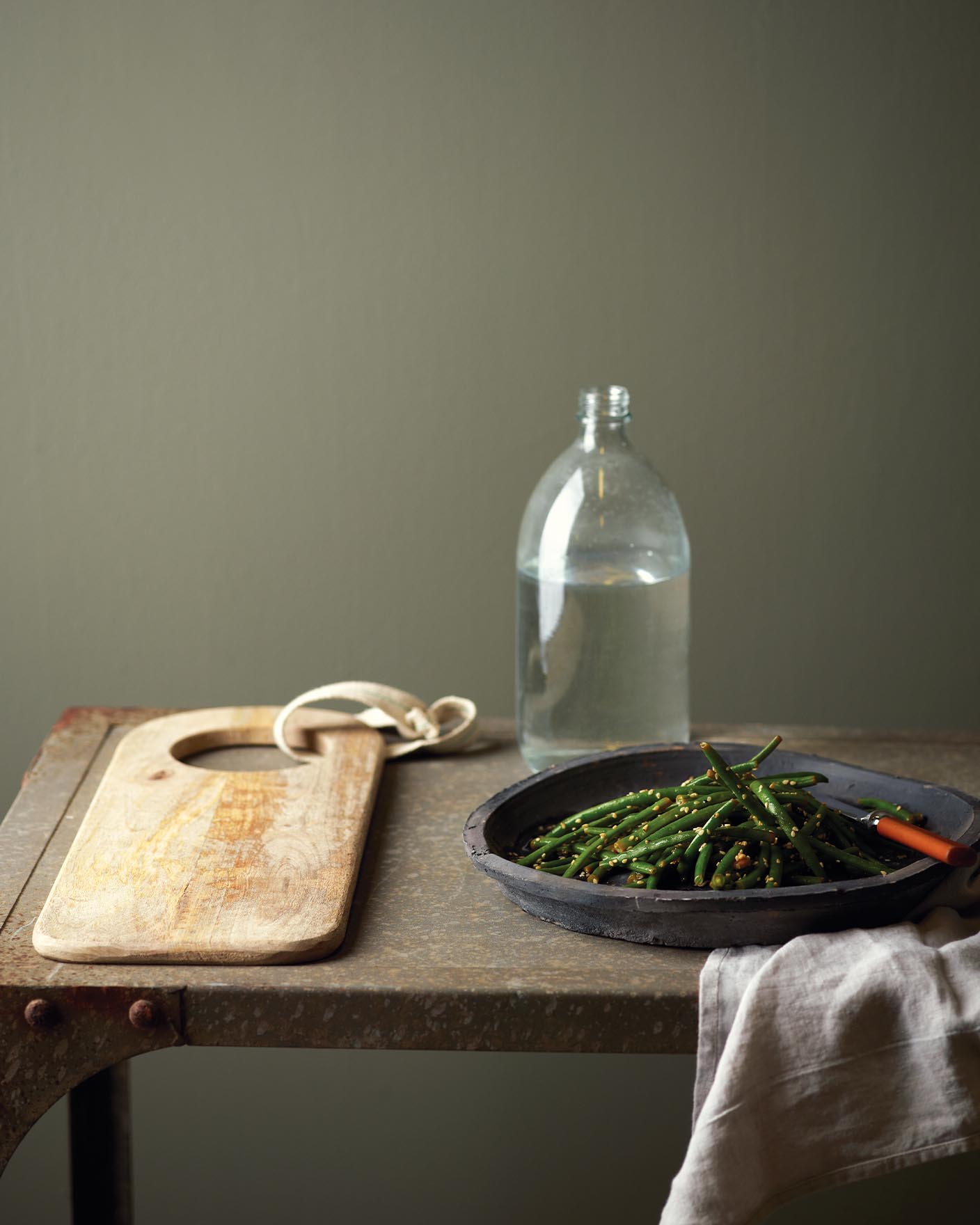 Grønne bønner serveres i en rustikk svart bolle på et forvitret metallbord, mot en mørk, dempet grønn vegg malt med Jotun 8304 Timian.
