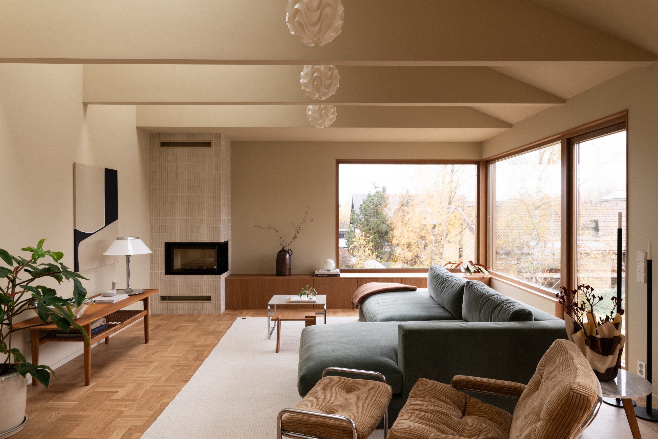 Stilren stue med moderne interiør der vegger og tak er malt med Jotun i den gulbeige fargen 11173 Humble Yellow, inkludert en grønn sofa, brun lenestol, peis og store vinduer som gir naturlig lys