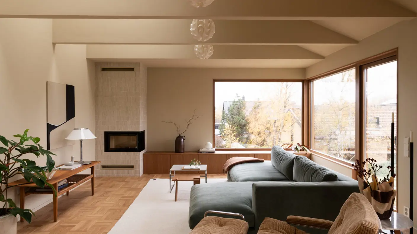 Stilren stue med moderne interiør der vegger og tak er malt med Jotun i den gulbeige fargen 11173 Humble Yellow, inkludert en grønn sofa, brun lenestol, peis og store vinduer som gir naturlig lys