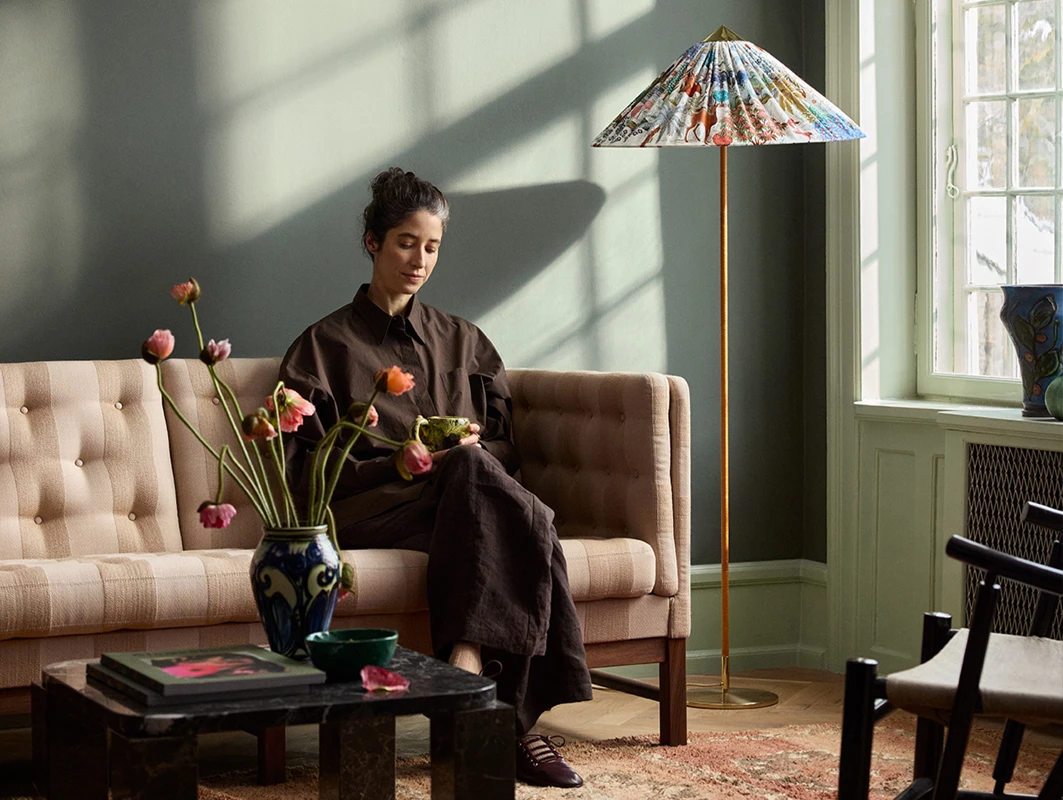Person som sitter på en sofa i en stilig stue med blomster i en vase på bordet og en fargerik gulvlampe i hjørnet, veggene er malt mørkegrønne i Jotun 7613 Northern Mystic