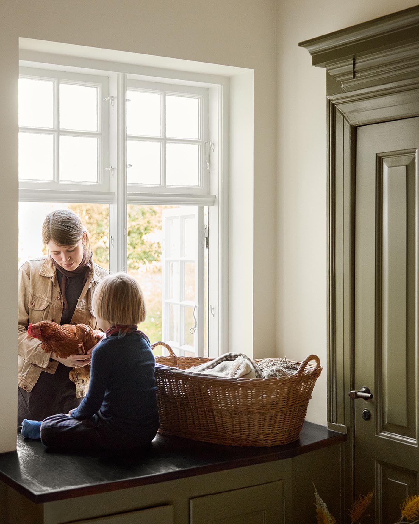 Woman and a child child on a kitchen counter by the window next to a olive-green cabinet in the colour 8597 Seaweed Green, against a muted yellow wall in the colour 12079 Gleam from Jotun.