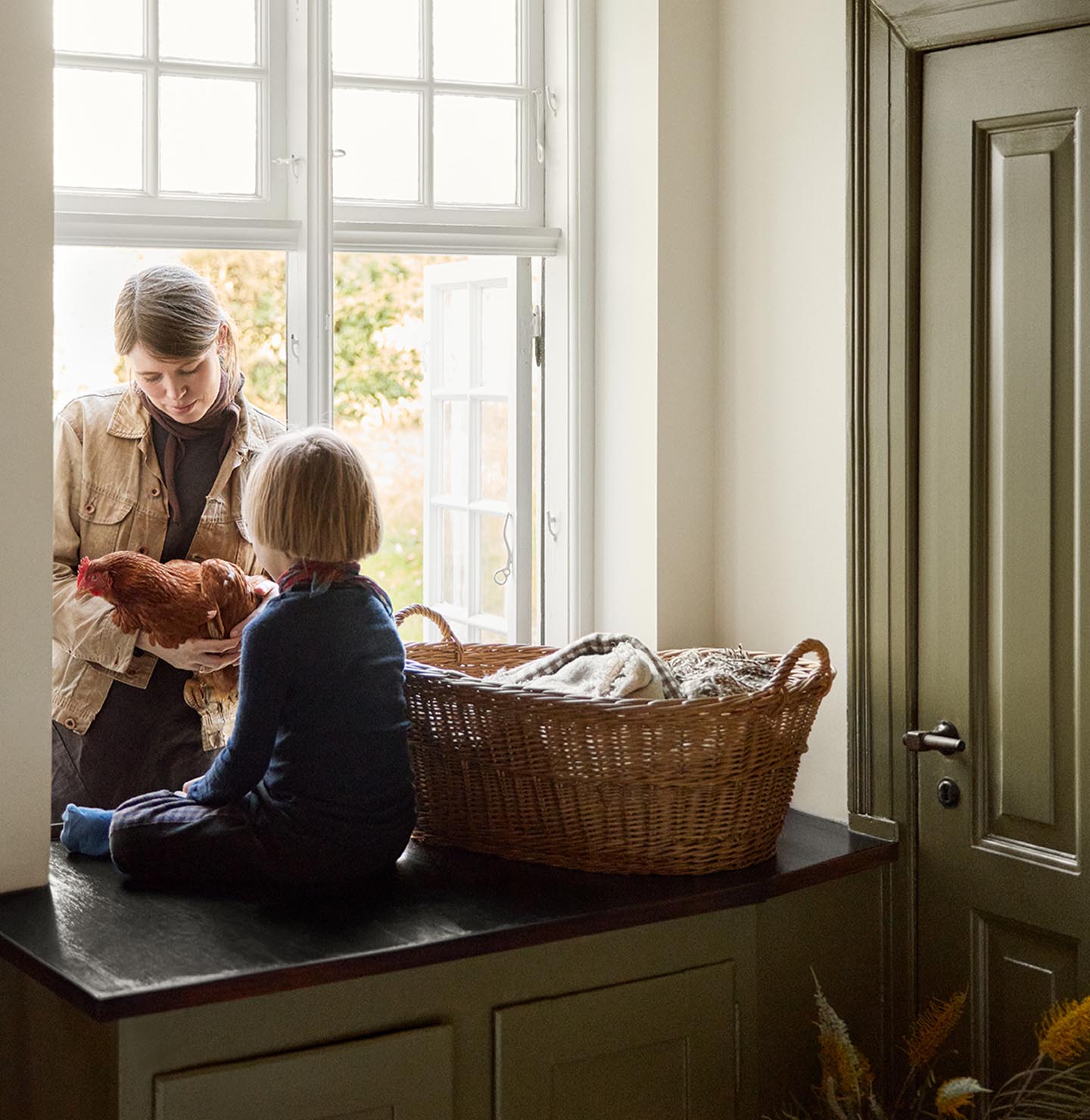 Woman and a child child on a kitchen counter by the window next to a olive-green cabinet in the colour 8597 Seaweed Green, against a muted yellow wall in the colour 12079 Gleam from Jotun.