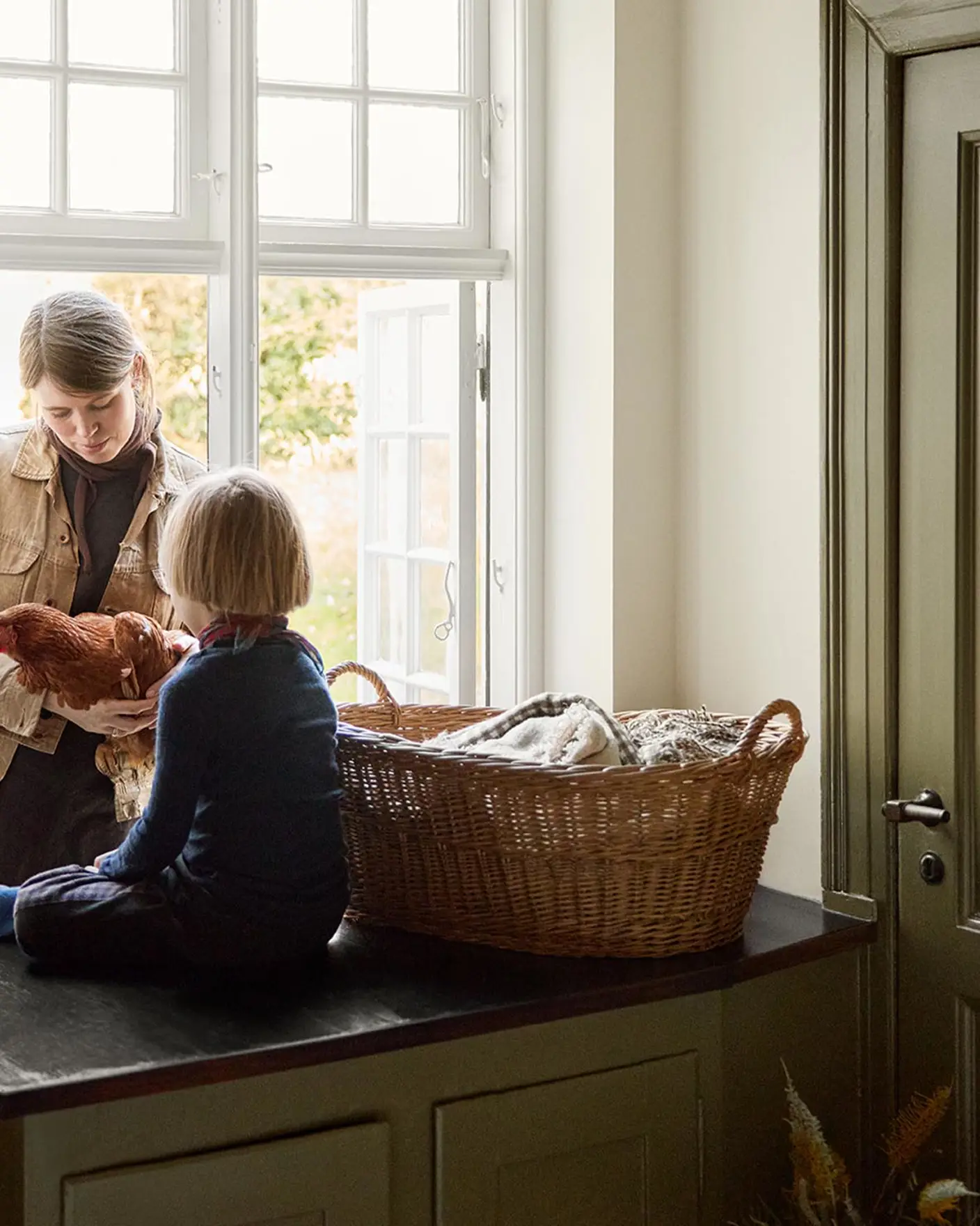 Woman and a child child on a kitchen counter by the window next to a olive-green cabinet in the colour 8597 Seaweed Green, against a muted yellow wall in the colour 12079 Gleam from Jotun.