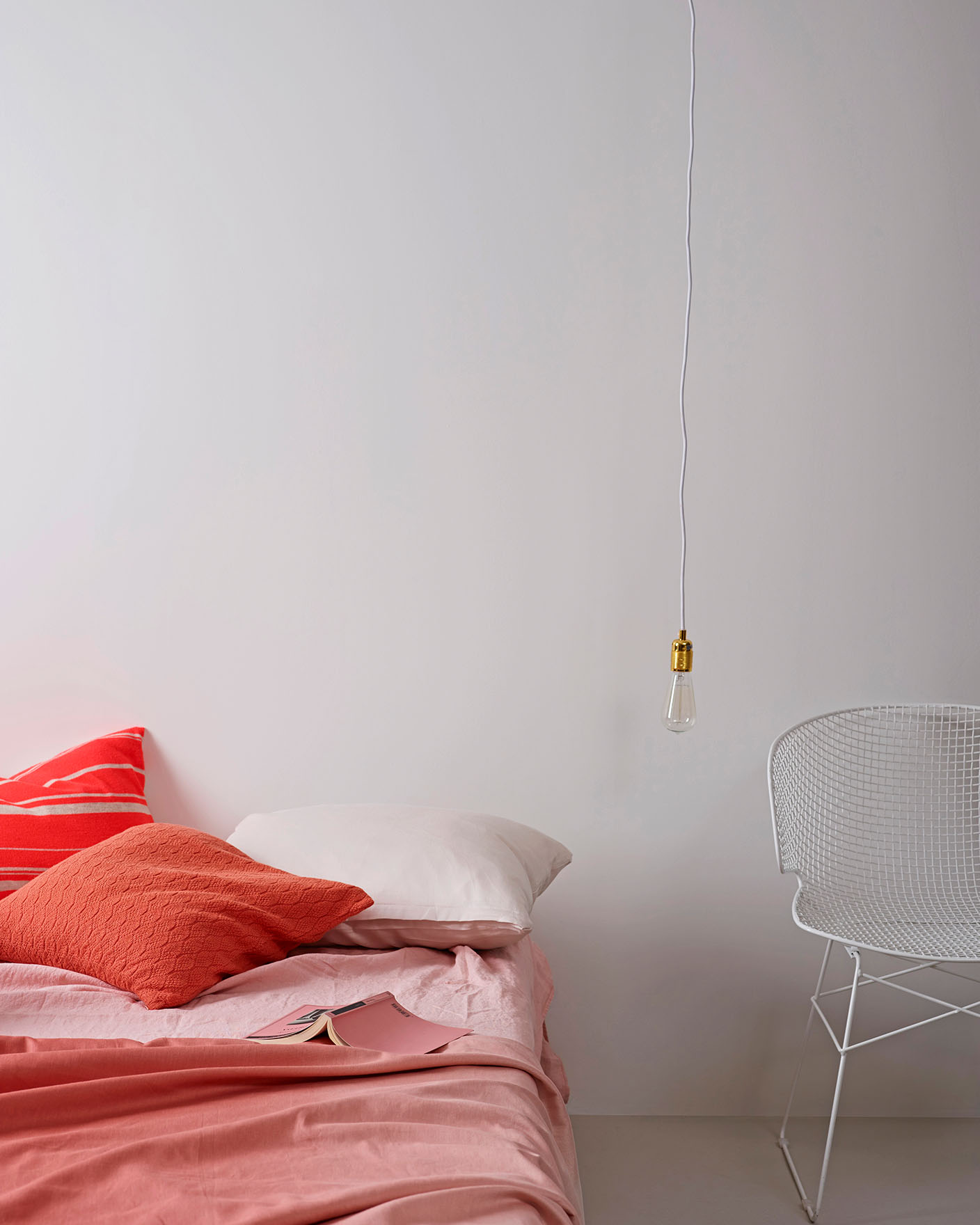 Mjukt rosa bäddset med korallfärgade accentkuddar på en låg säng, kombinerat med en vit trådstol mot en ljus, gyllene vit vägg målad med Jotun 1624 Lätthet.