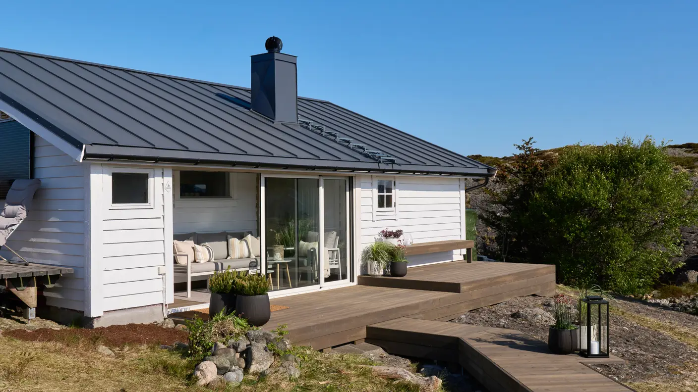 Hvit hytte med moderne design og terrasse, omgitt av natur og blå himmel, ideell for sommerferie i Norge