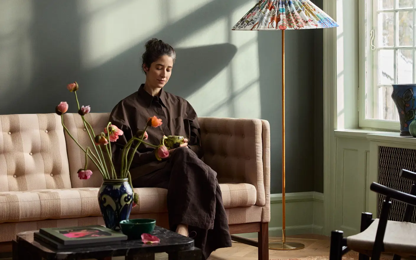 En kvinne som sitter på en beige sofa, holder en kopp, i en solfylt grønnmalt stue i Jotun Northern Mystic, med en fargerik gulvlampe og en blomster vase på et salongbord