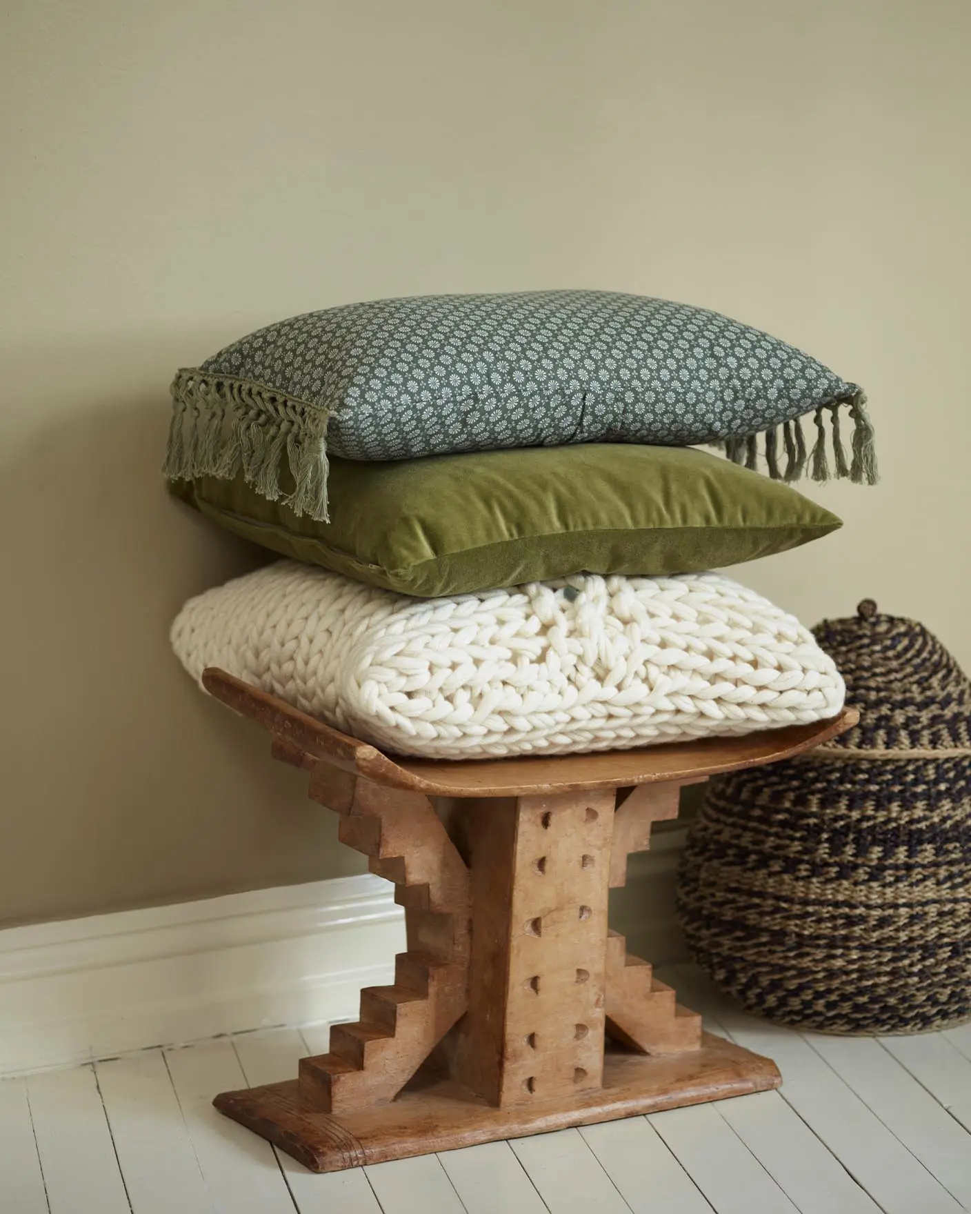 Stack of textured cushions on a carved wooden stool, with a woven basket beside it, set against a muted, beige yellow wall painted with Jotun 1938 Tea Leaves.