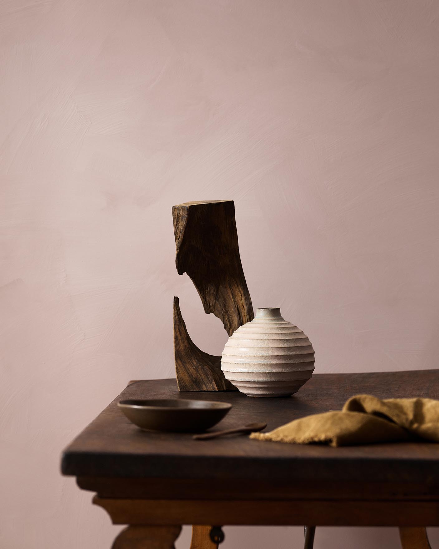 Wooden table with a ceramic vase, sculpted wood, and a dark bowl against a muted pink background painted with LADY Minerals in the colour 20054 Silky Pink from Jotun.