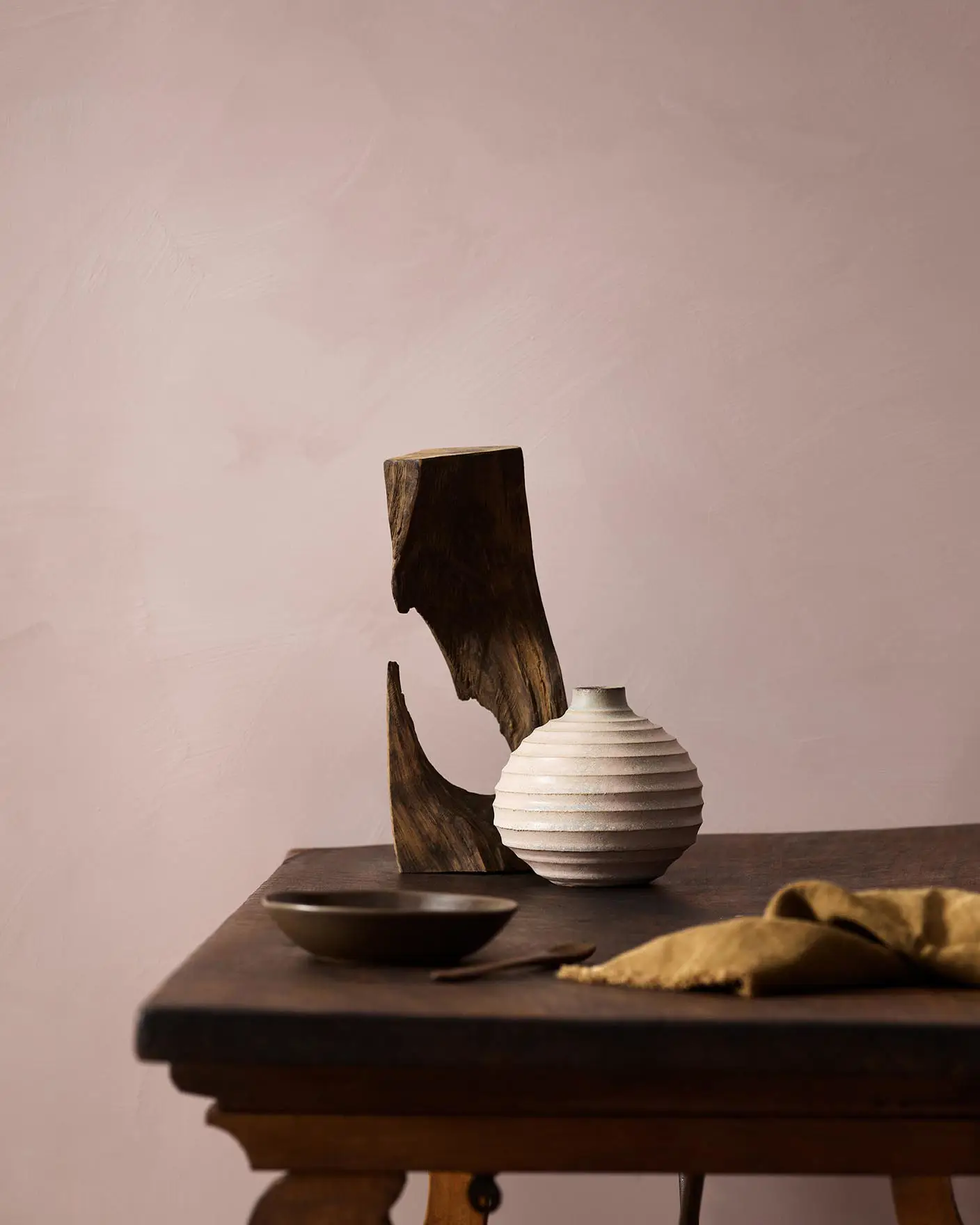 Wooden table with a ceramic vase, sculpted wood, and a dark bowl against a muted pink background painted with LADY Minerals in the colour 20054 Silky Pink from Jotun.