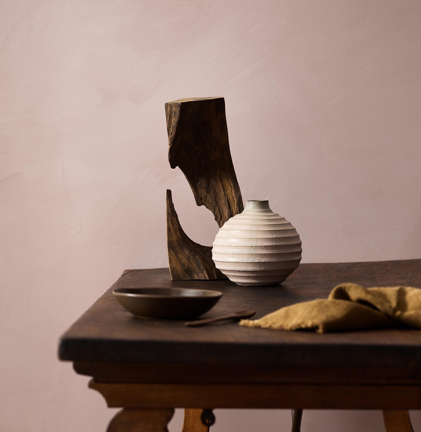 Wooden table with a ceramic vase, sculpted wood, and a dark bowl against a muted pink background painted with LADY Minerals in the colour 20054 Silky Pink from Jotun.