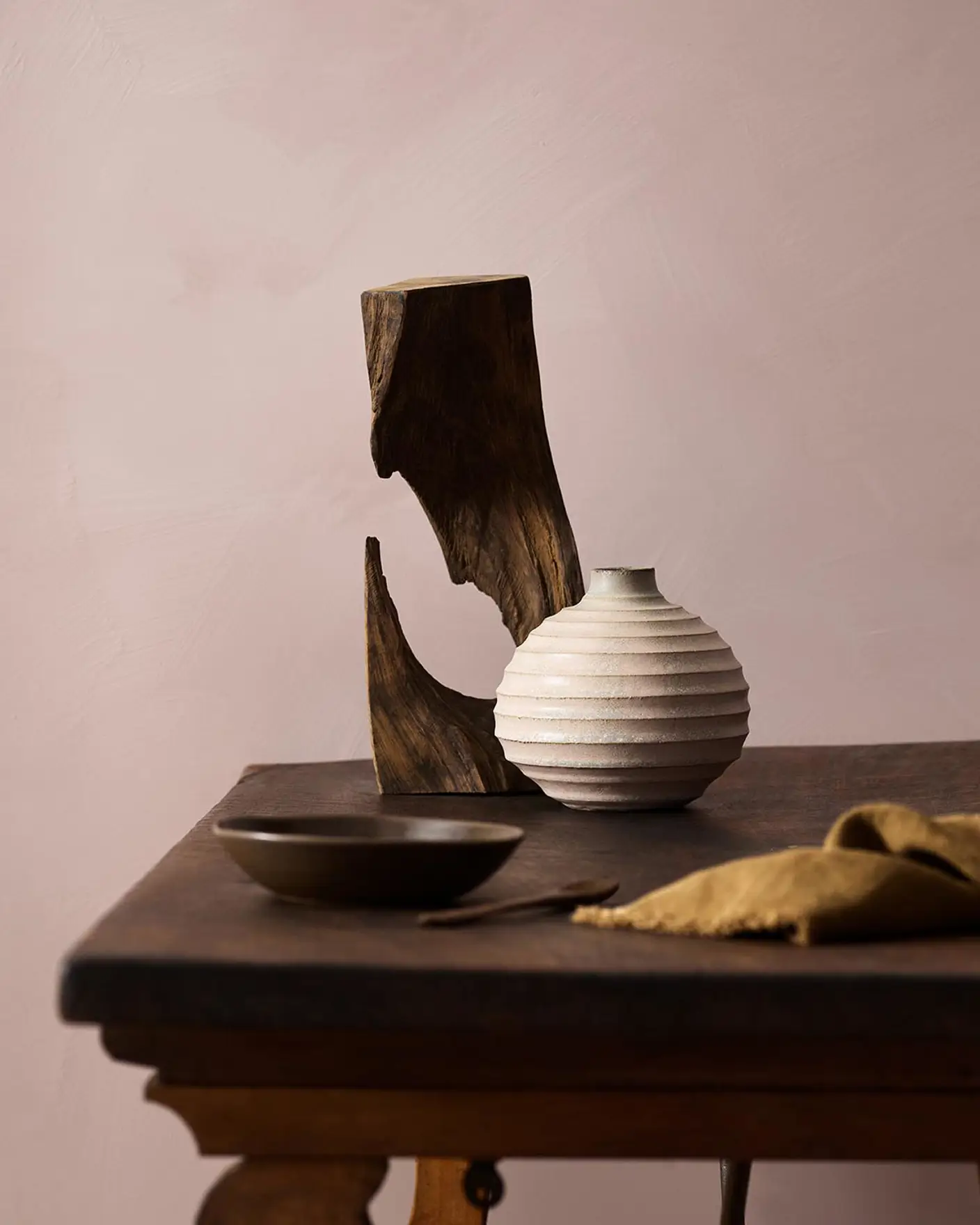 Wooden table with a ceramic vase, sculpted wood, and a dark bowl against a muted pink background painted with LADY Minerals in the colour 20054 Silky Pink from Jotun.