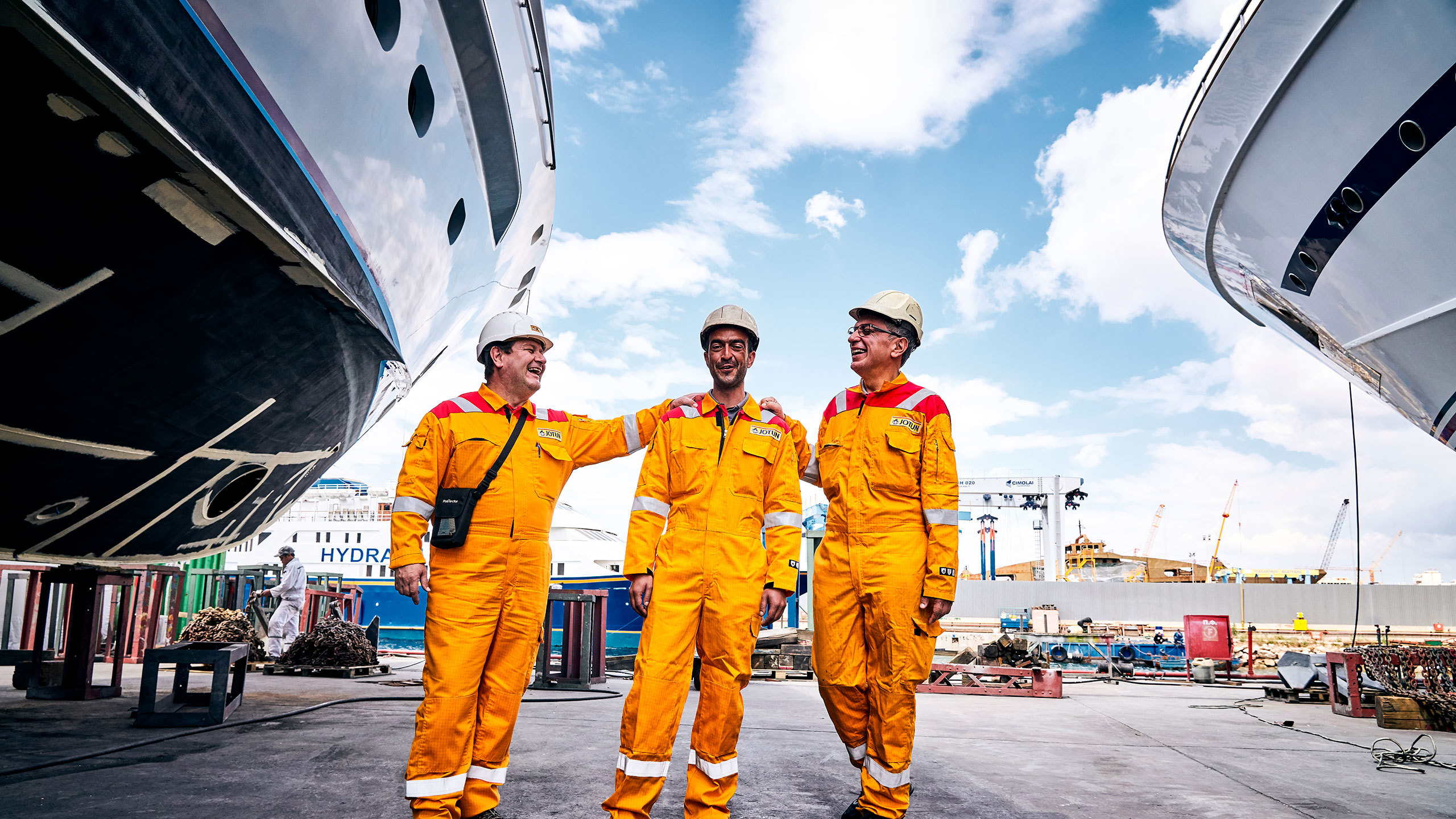 Tre glada arbetare i orange arbetskläder och hjälmar står mellan två stora båtar på ett varv en solig dag