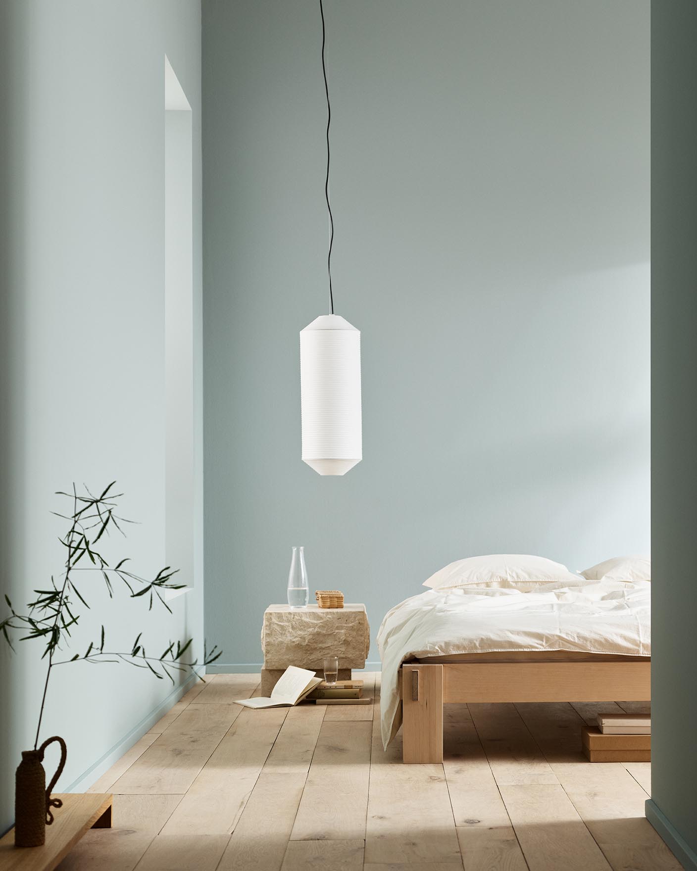 Minimalist bedroom with white bedding, a stone side table, and a plant against muted blue walls painted with the colour 6378 Iconic from Jotun.