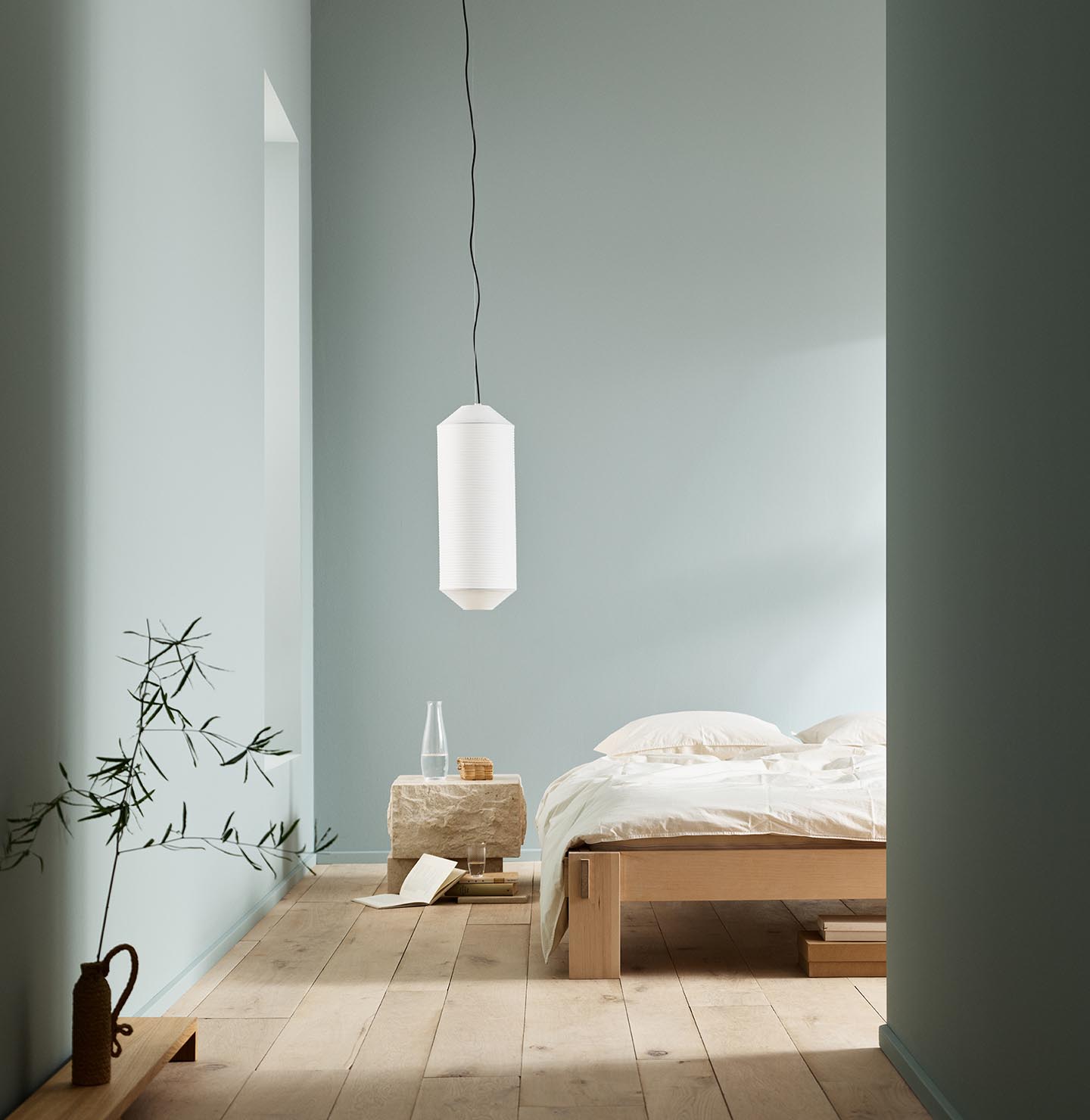 Minimalist bedroom with white bedding, a stone side table, and a plant against muted blue walls painted with the colour 6378 Iconic from Jotun.