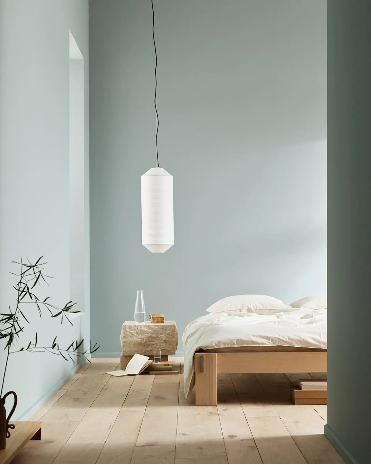 Minimalist bedroom with white bedding, a stone side table, and a plant against muted blue walls painted with the colour 6378 Iconic from Jotun.