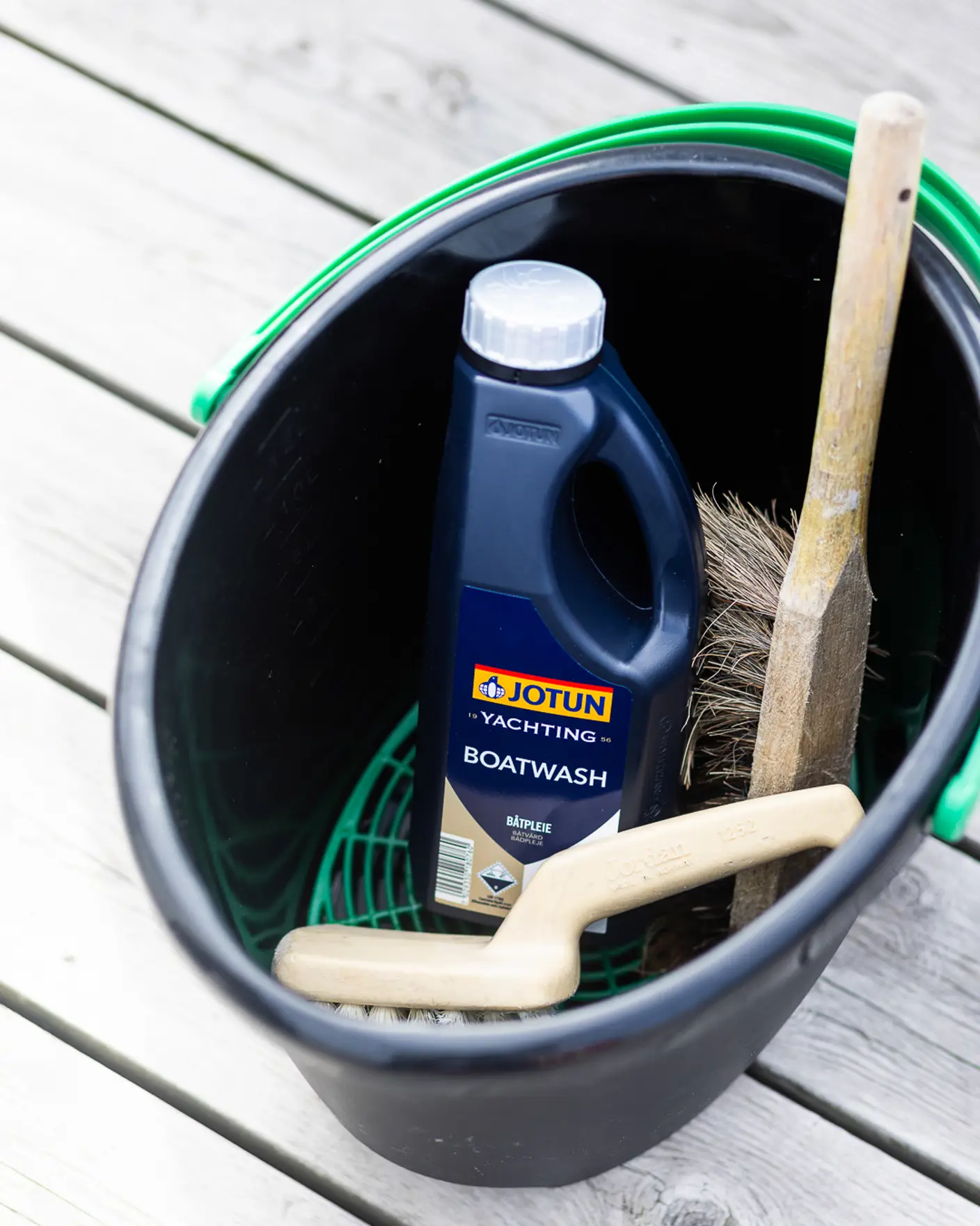 Emballasje med Jotun Boatwash, kost og børste i en bøtte på brygge med vann i bakgrunnen.
