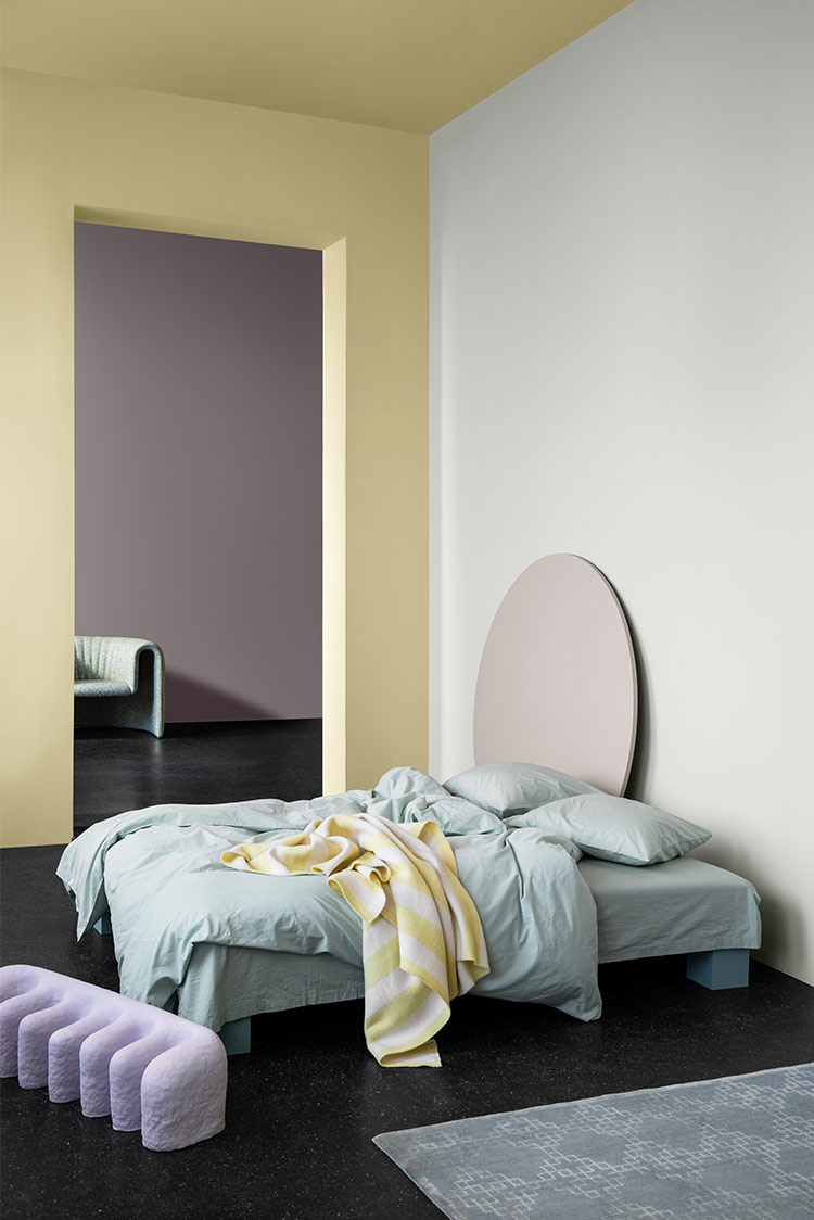 Moderne soveromsinteriør med et fargeskjema i pastell, med en lys gul vegg i Jotun 1775 Fresh Pasta, en hvit vegg i Jotun 7236 Chi, en lav seng med lys salviefarget sengetøy, et gult og hvitt stripete teppe og et stort rundt hodegjerde i lys lilla Jotun 3377 Slate Lavender. En lilla polstret benk og et gulvteppe kompletterer det moderne uttrykket