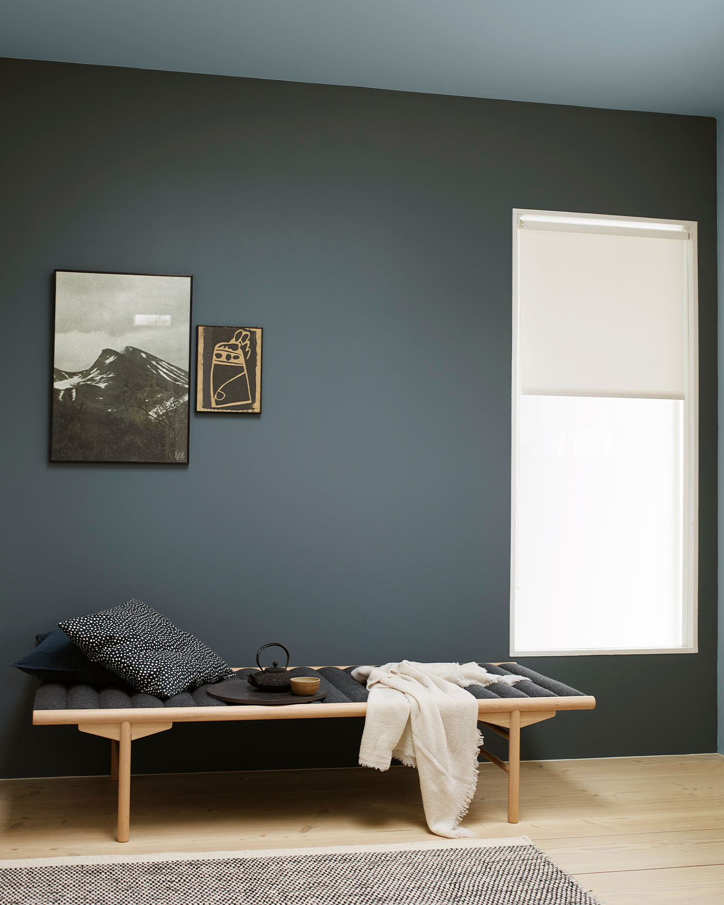 Serene space with a wooden bench layered with dark cushions and a cream throw, set against a muted, greenish blue wall painted with Jotun 5030 St. Paul Blue. Ceiling painted with Jotun 5249 Artic Grey, a muted greenish blue.