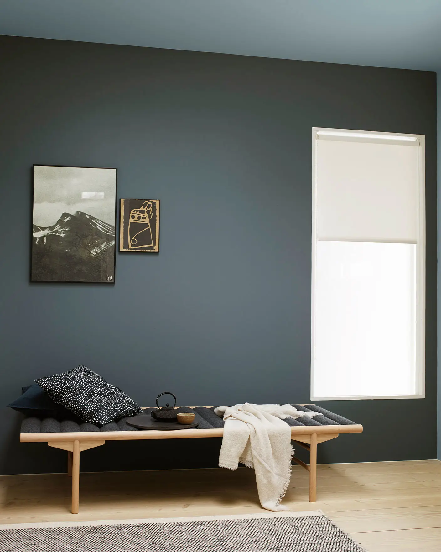 Serene space with a wooden bench layered with dark cushions and a cream throw, set against a muted, greenish blue wall painted with Jotun 5030 St. Paul Blue. Ceiling painted with Jotun 5249 Artic Grey, a muted greenish blue.