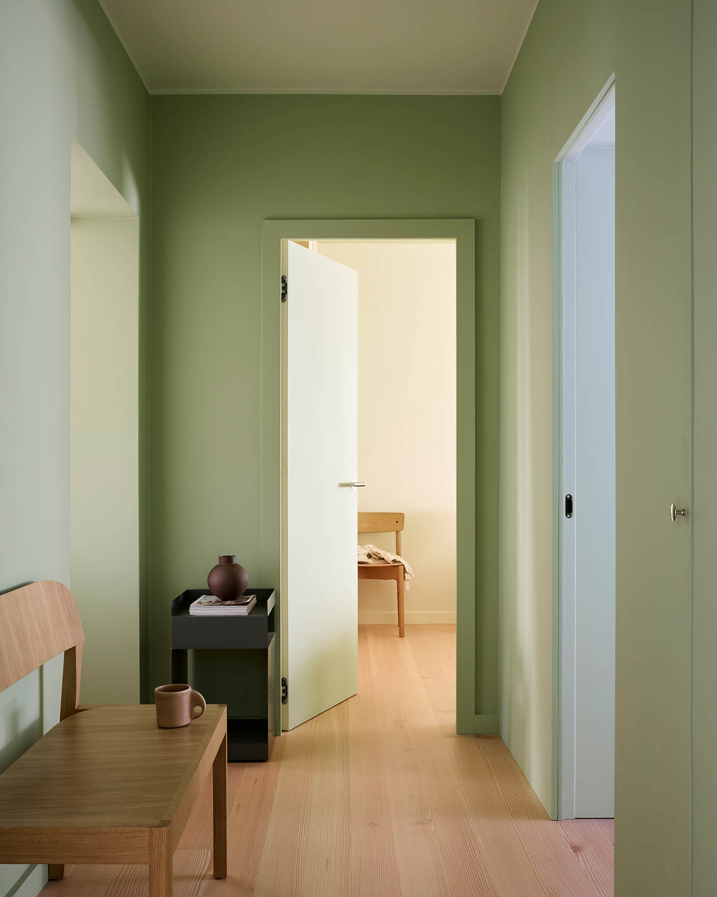 Hallway painted with Jotun 0470 Avocado Green, 8124 Malmoe, 7718 Secret Green, and 6167 Spring Breeze. White door opens to beige room with wooden furniture