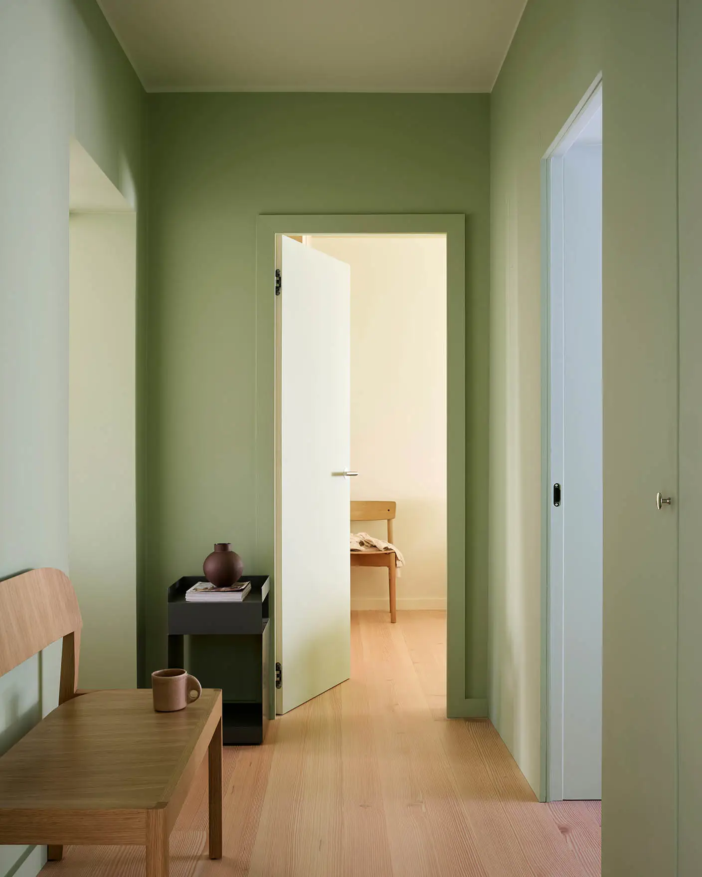 Hallway painted with Jotun 0470 Avocado Green, 8124 Malmoe, 7718 Secret Green, and 6167 Spring Breeze. White door opens to beige room with wooden furniture