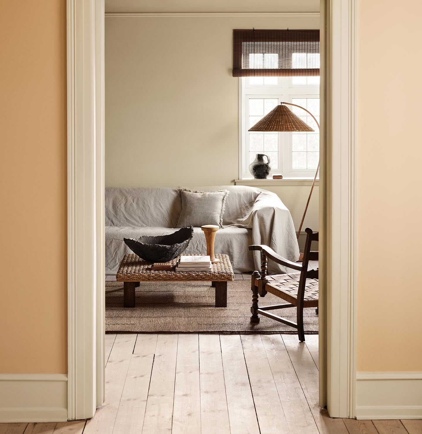 Living room with a light sofa, woven coffee table against a muted yellow wall painted in 12080 Soft Radiance. Wall in front in the colour 11202 Mild Ochre.