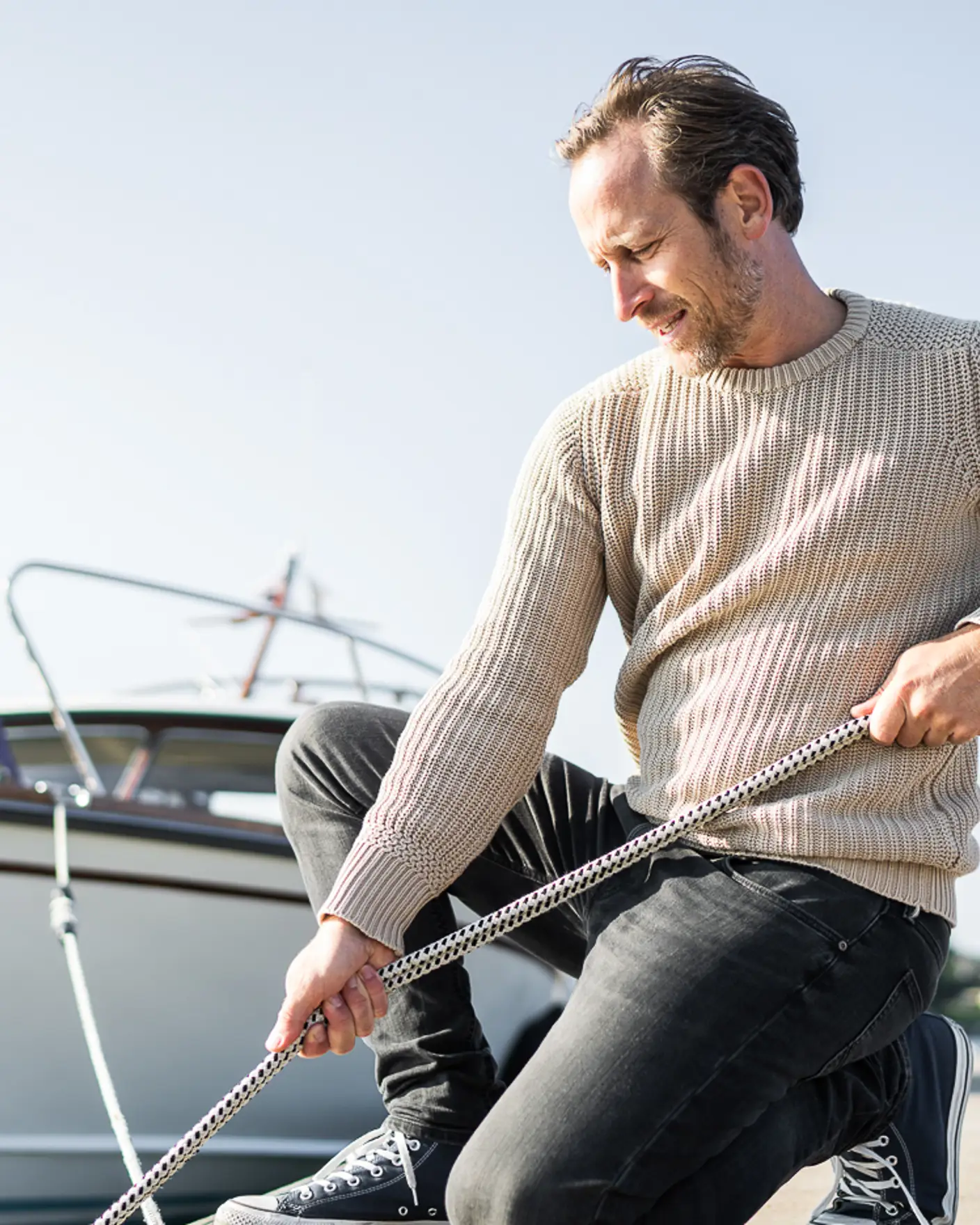 Mann drar i tau på bryggekant med båt og blå himmel i bakgrunnen.