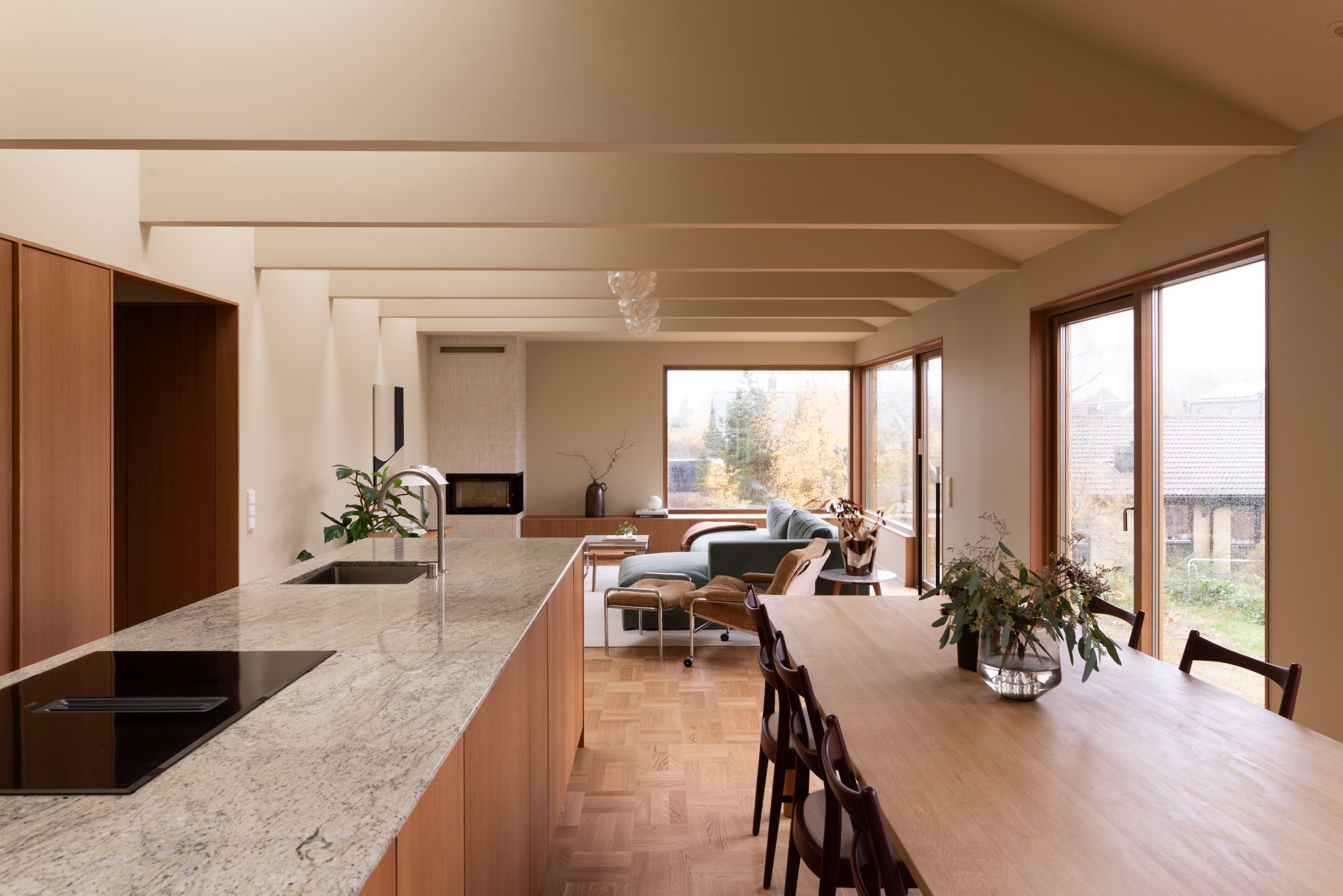 Moderne kjøkken og stue med åpen planløsning, preget av store vinduer som slipper inn naturlig lys. Rommet har en kjøkkenøy i marmor, en tre-spisebord, og en komfortabel sittegruppe med utsikt mot hagen, vegger og tak malt i Jotun 11173 Humble Yellow