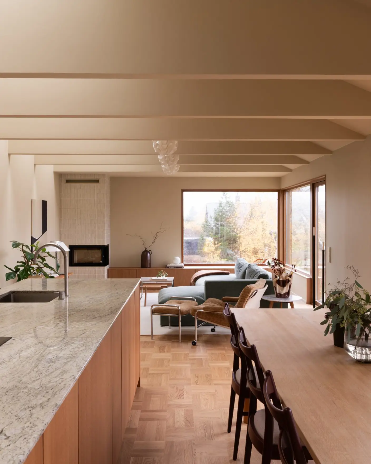 Moderne kjøkken og stue med åpen planløsning, preget av store vinduer som slipper inn naturlig lys. Rommet har en kjøkkenøy i marmor, en tre-spisebord, og en komfortabel sittegruppe med utsikt mot hagen, vegger og tak malt i Jotun 11173 Humble Yellow