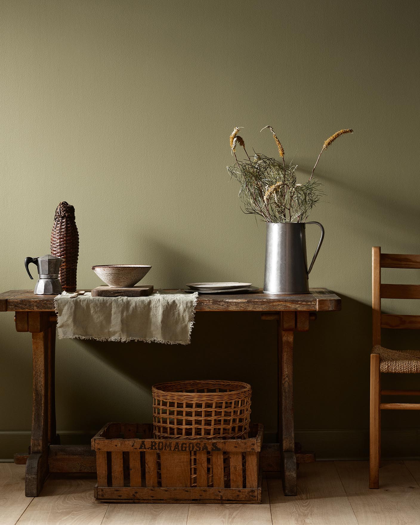 Rustic wooden table with vintage kitchenware and a wicker basket against a olivegreen wall painted with the colour 8597 Seaweed Green from Jotun.