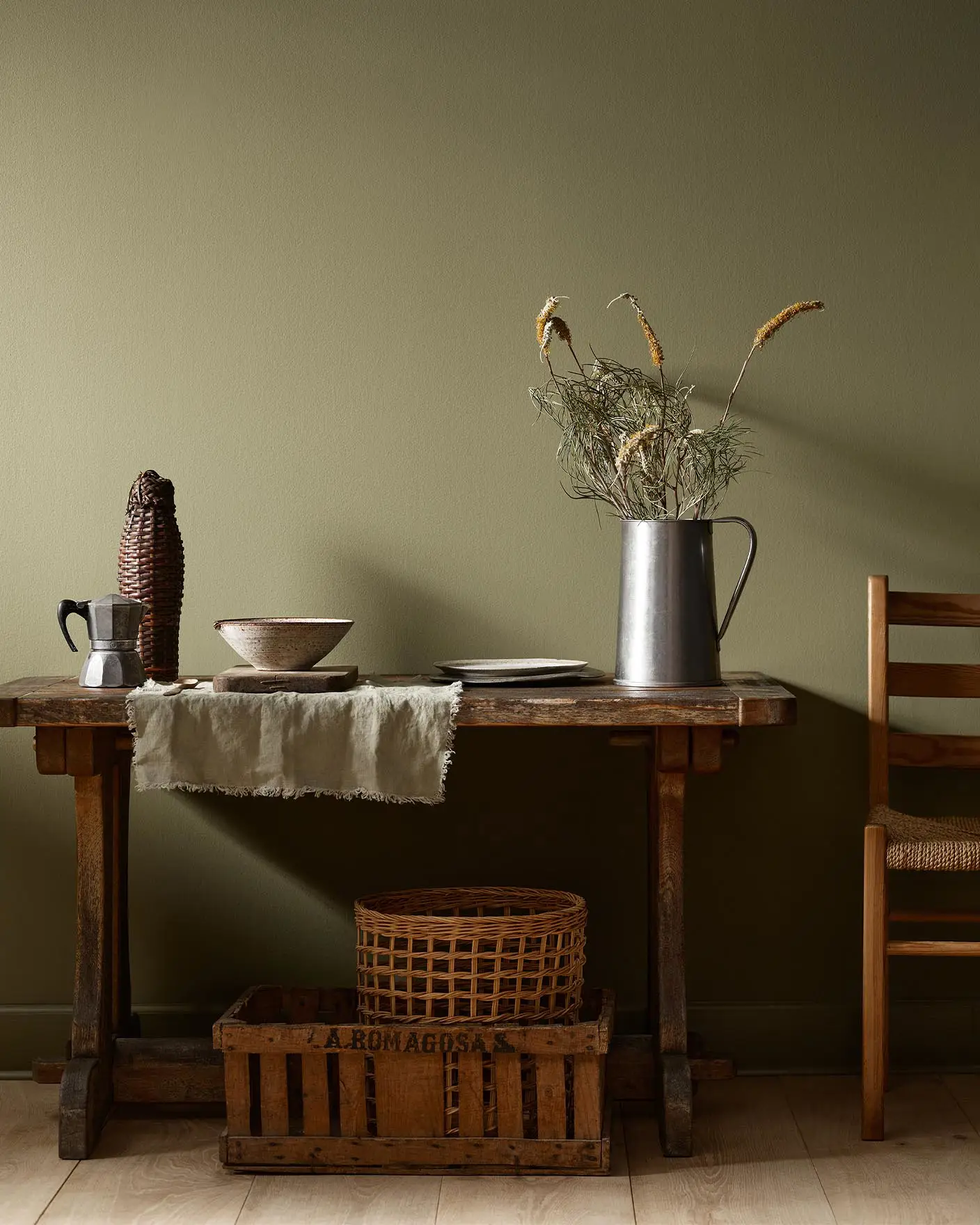 Rustic wooden table with vintage kitchenware and a wicker basket against a olivegreen wall painted with the colour 8597 Seaweed Green from Jotun.