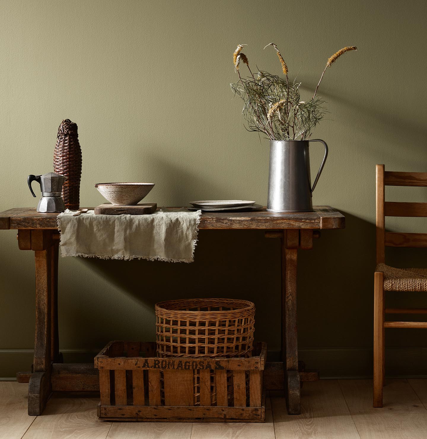 Rustic wooden table with vintage kitchenware and a wicker basket against a olivegreen wall painted with the colour 8597 Seaweed Green from Jotun.