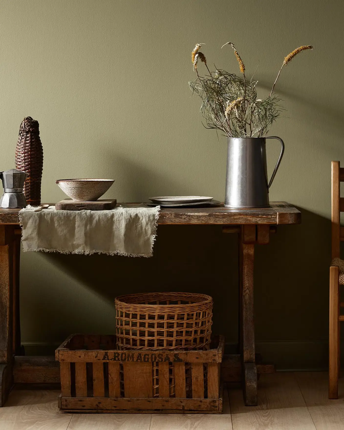 Rustic wooden table with vintage kitchenware and a wicker basket against a olivegreen wall painted with the colour 8597 Seaweed Green from Jotun.