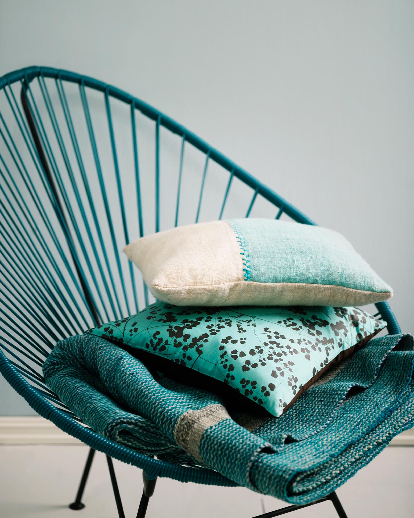 Teal woven chair holding a stack of textured cushions and a blanket in turquoise and cream tones, set against a muted, greenish blue wall painted with Jotun 5225 Lake View.