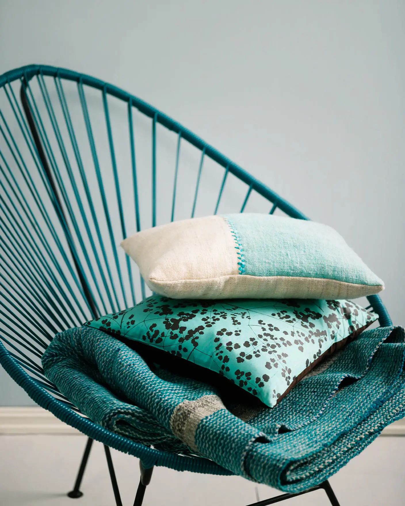 Teal woven chair holding a stack of textured cushions and a blanket in turquoise and cream tones, set against a muted, greenish blue wall painted with Jotun 5225 Lake View.