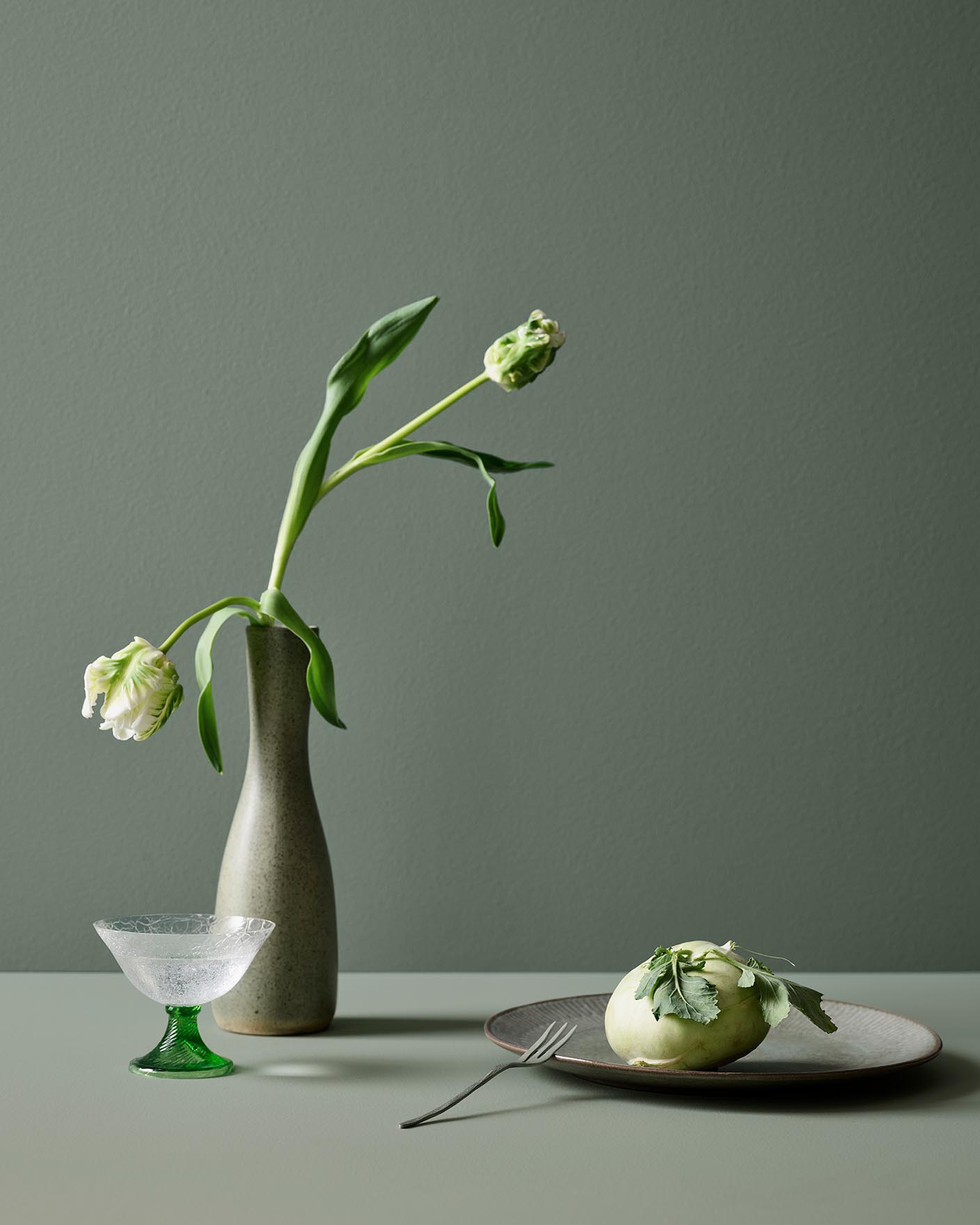 A tulip in a vase on a table of soft green tones painted with Jotun 7685 Subtle Green, with a background wall painted with 7686 Mindful Green from Jotun.