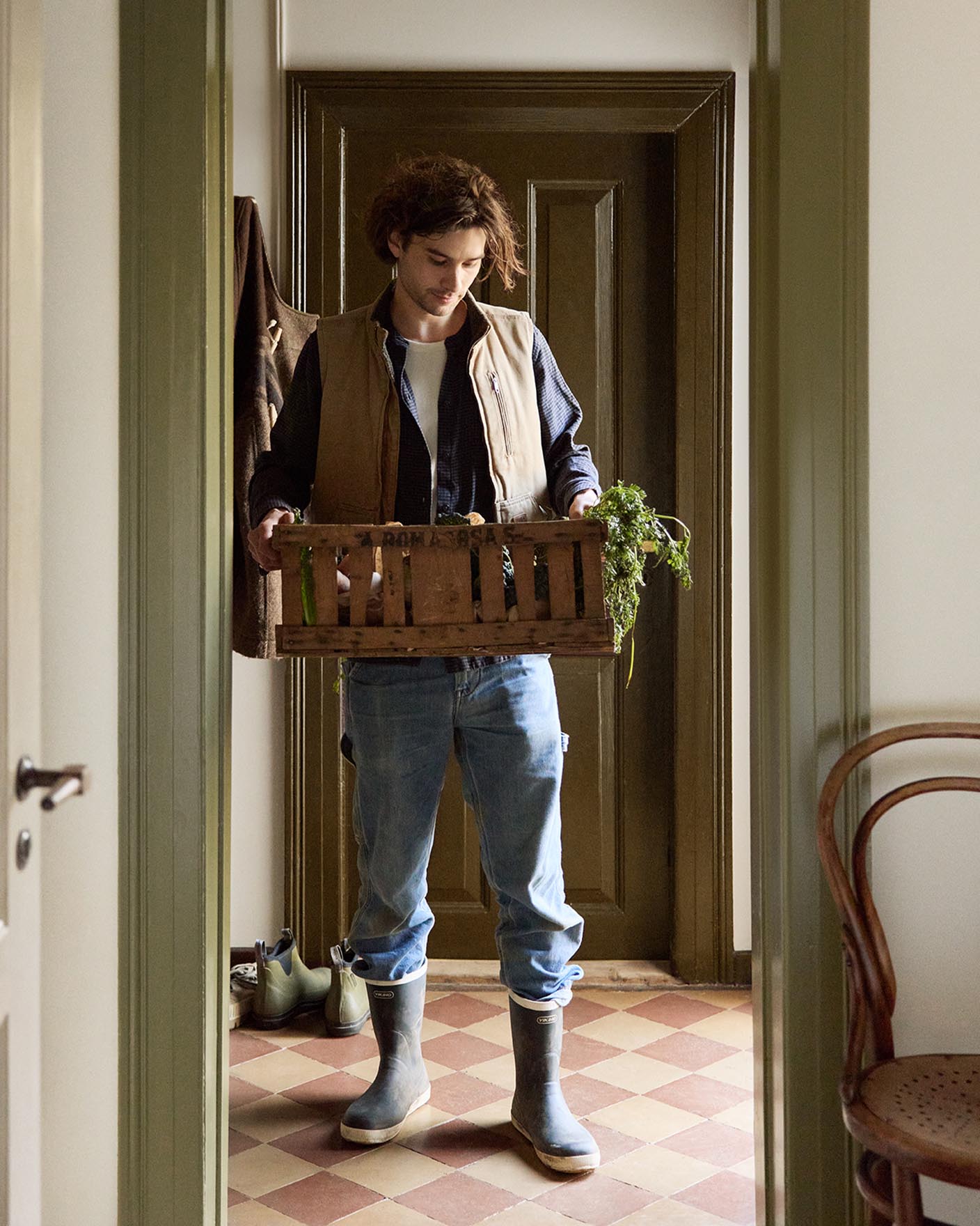 Person in a doorway holding a crate with greenery. Walls painted in the muted yellow colour 12079 Gleam from Jotun. Olive-green trims in Jotun's 8597 Seawood Green.