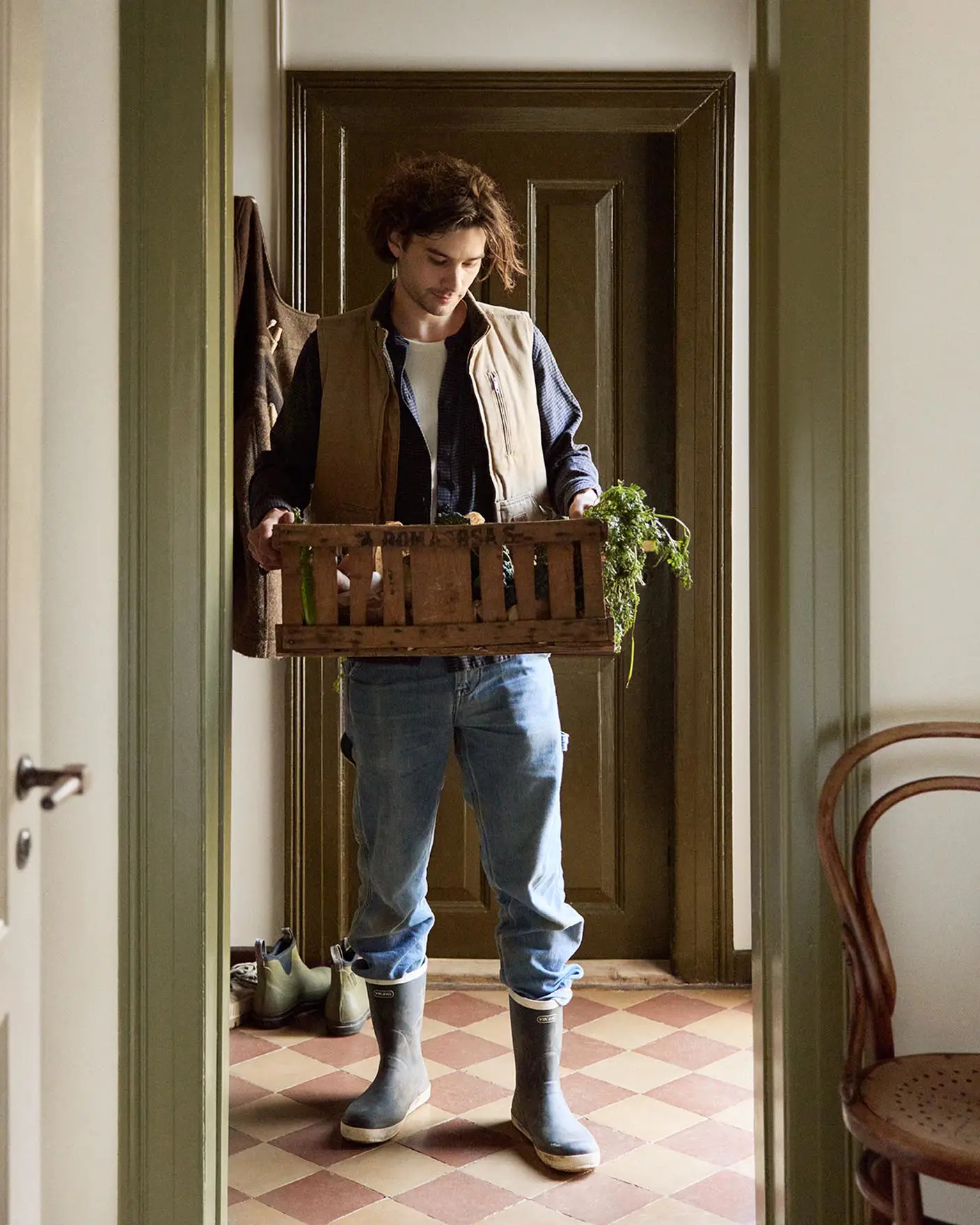 Person in a doorway holding a crate with greenery. Walls painted in the muted yellow colour 12079 Gleam from Jotun. Olive-green trims in Jotun's 8597 Seawood Green.