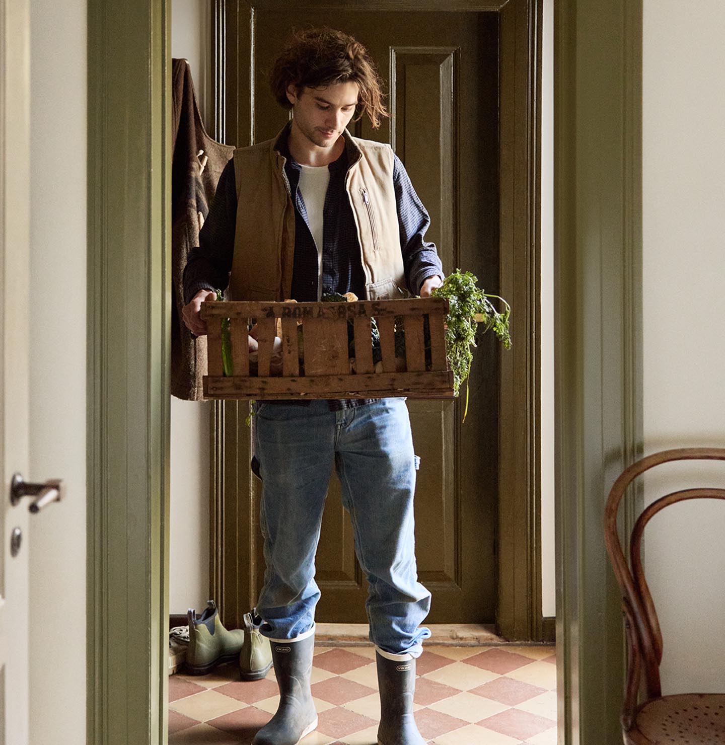 Person in a doorway holding a crate with greenery. Walls painted in the muted yellow colour 12079 Gleam from Jotun. Olive-green trims in Jotun's 8597 Seawood Green.