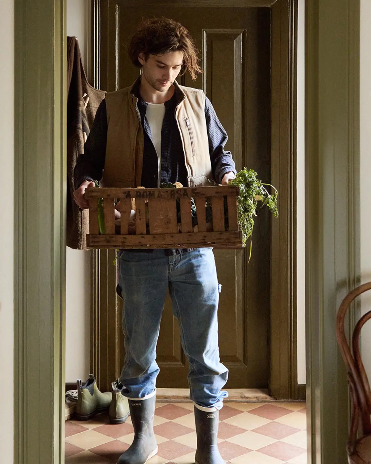 Person in a doorway holding a crate with greenery. Walls painted in the muted yellow colour 12079 Gleam from Jotun. Olive-green trims in Jotun's 8597 Seawood Green.