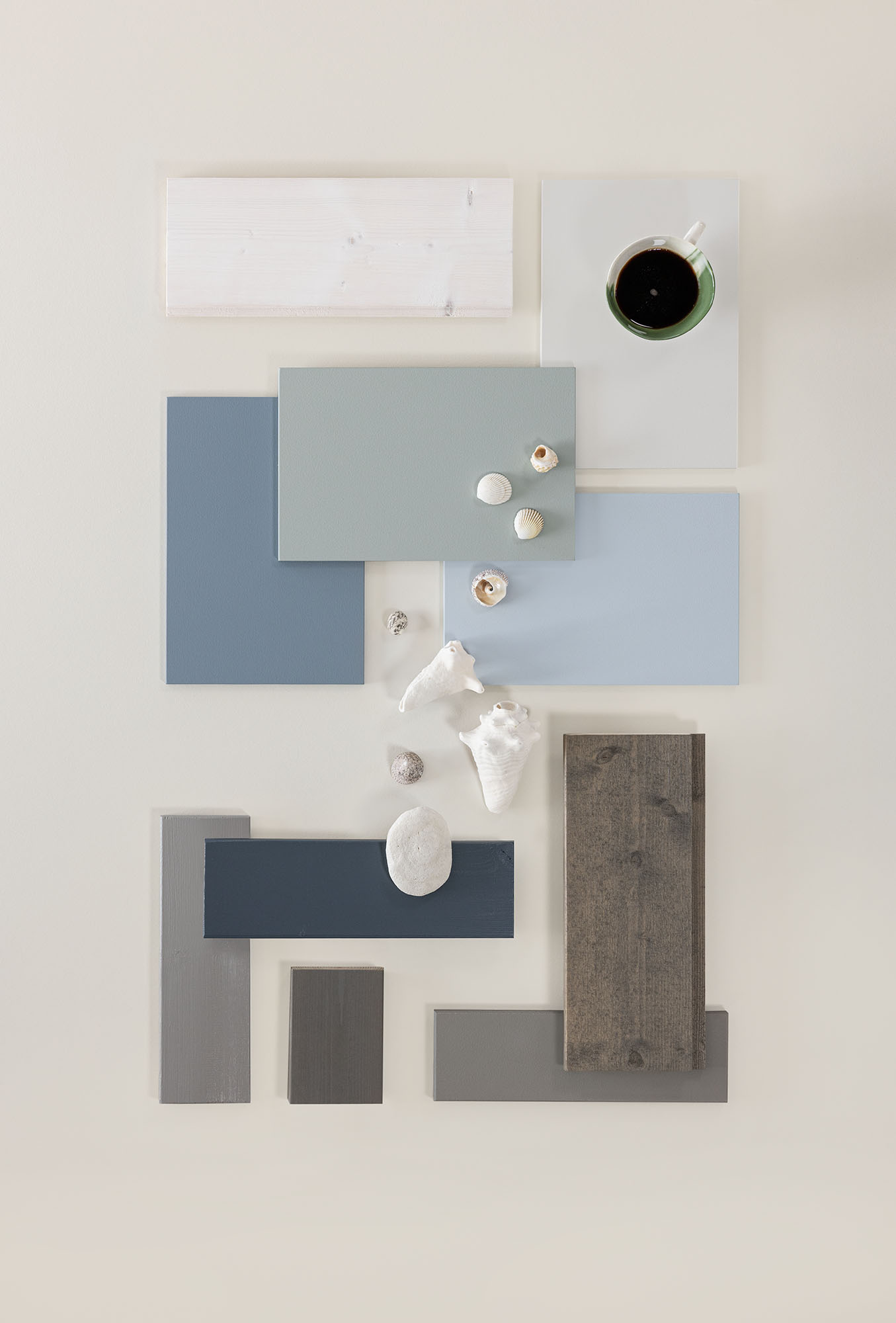 Interiørdesigninspirert flatlay med blå og grå fargetoner, ulike teksturer av malingprøver, treverk og små skjell, samt en kopp kaffe