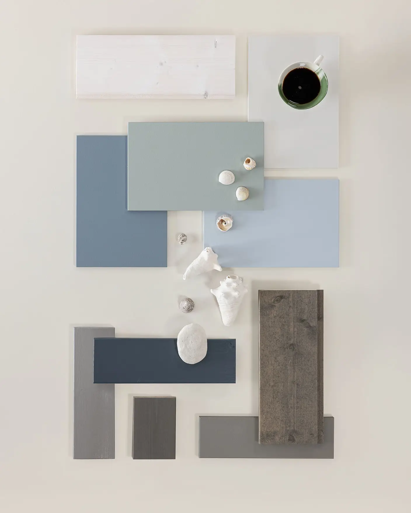 Interiørdesigninspirert flatlay med blå og grå fargetoner, ulike teksturer av malingprøver, treverk og små skjell, samt en kopp kaffe