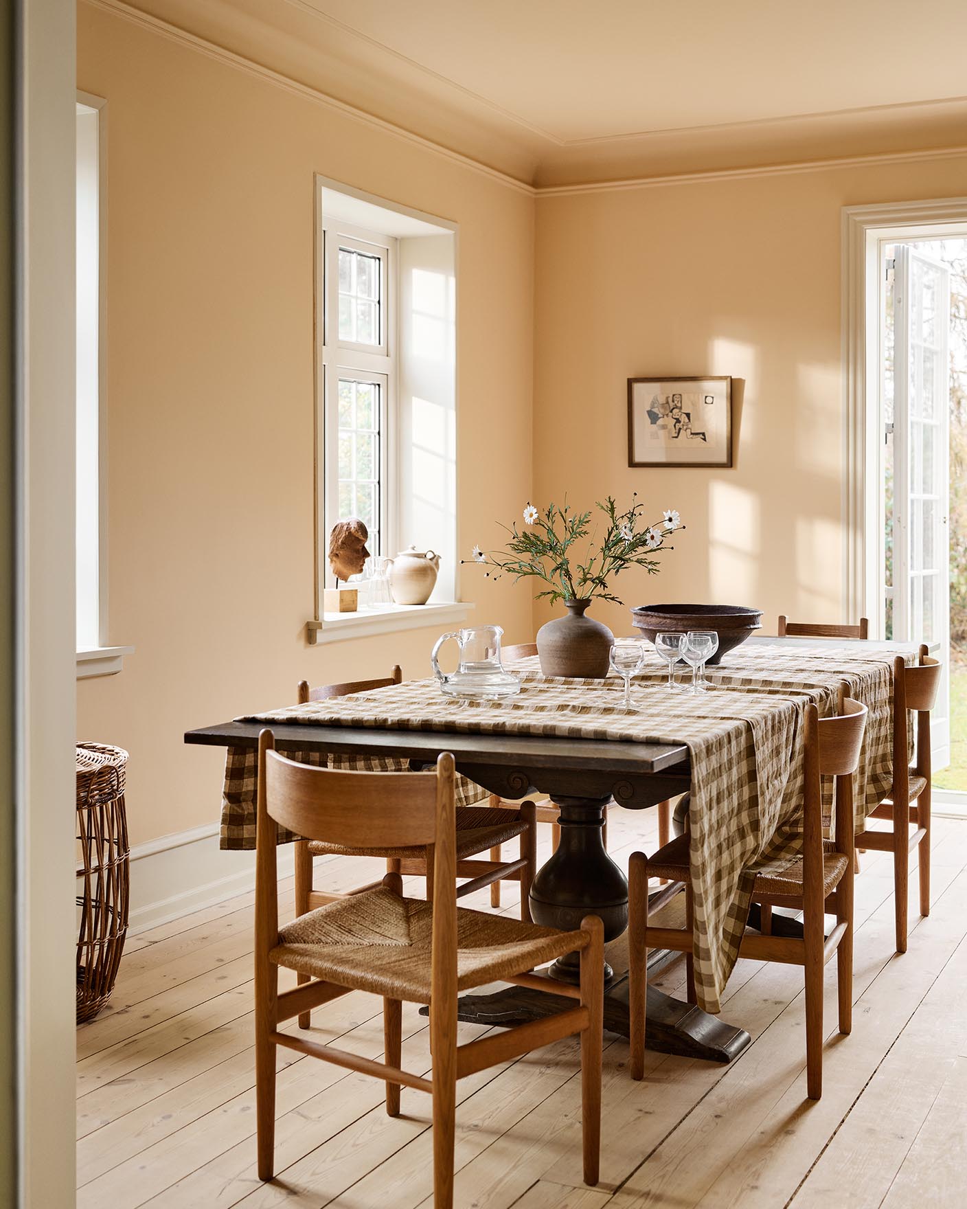 Dining room with a checkered tablecloth, wooden chairs, against an ochre yellow wall painted in the colour 11202 Mild Ochre from Jotun.