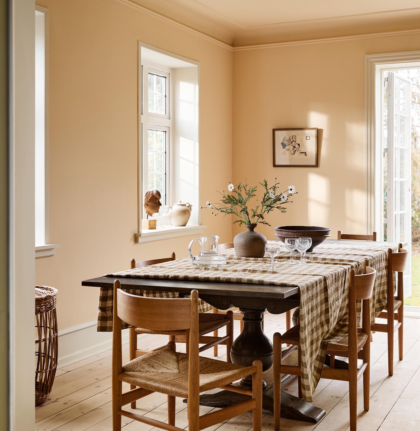 Dining room with a checkered tablecloth, wooden chairs, against an ochre yellow wall painted in the colour 11202 Mild Ochre from Jotun.