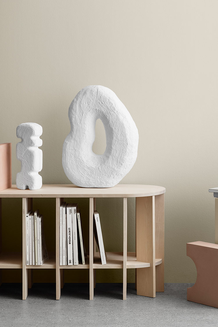 Moderne minimalistisk interiør med abstrakte skulpturer på beige hyller, bøker organisert vertikalt, og naturlig lys på en greige vegg malt i Jotun 12182 Gente Whisper