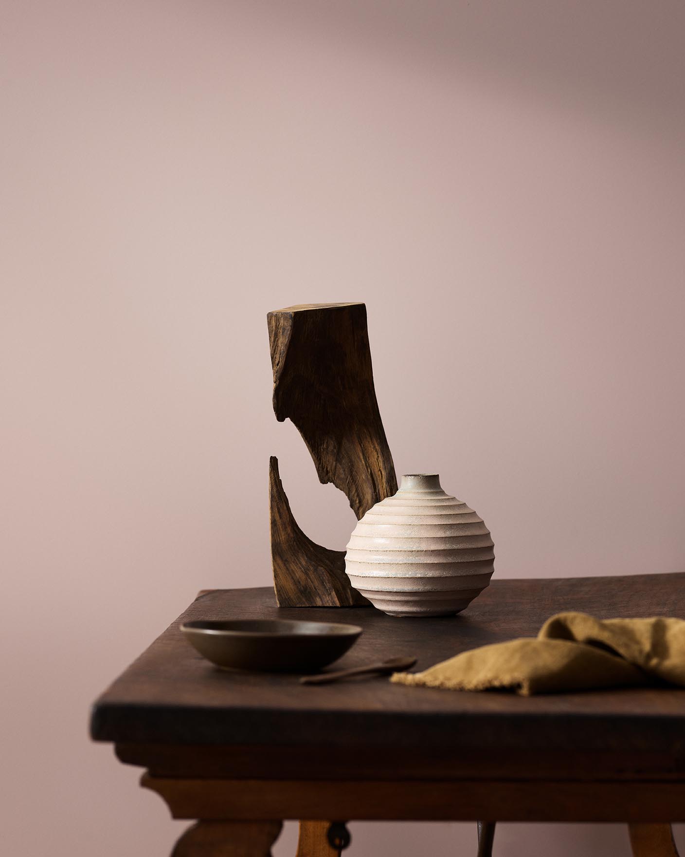Wooden table with a ceramic vase, sculpted wood, and a dark bowl against a muted pink background painted with the colour 20054 Silky Pink from Jotun.
