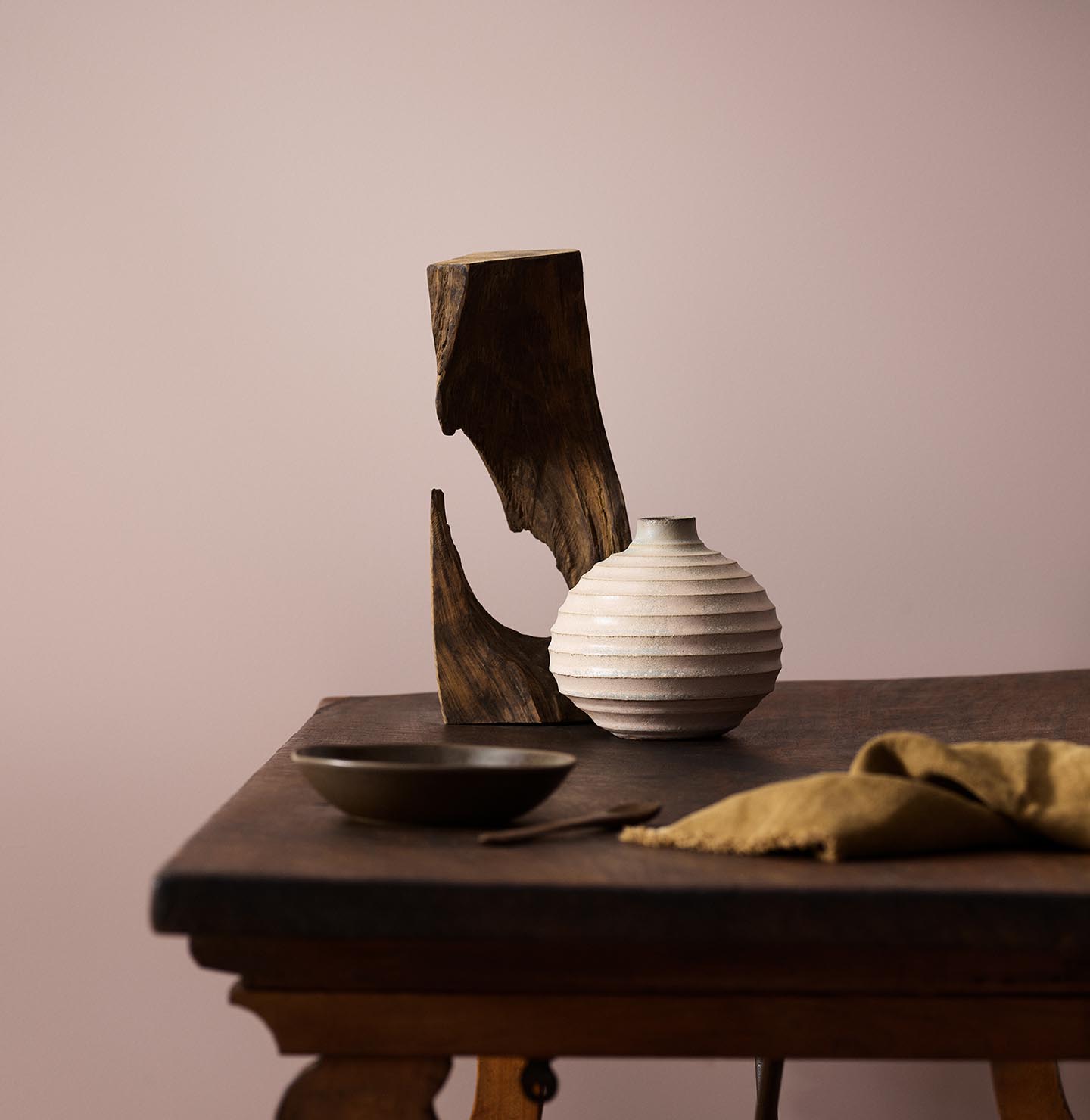 Wooden table with a ceramic vase, sculpted wood, and a dark bowl against a muted pink background painted with the colour 20054 Silky Pink from Jotun.