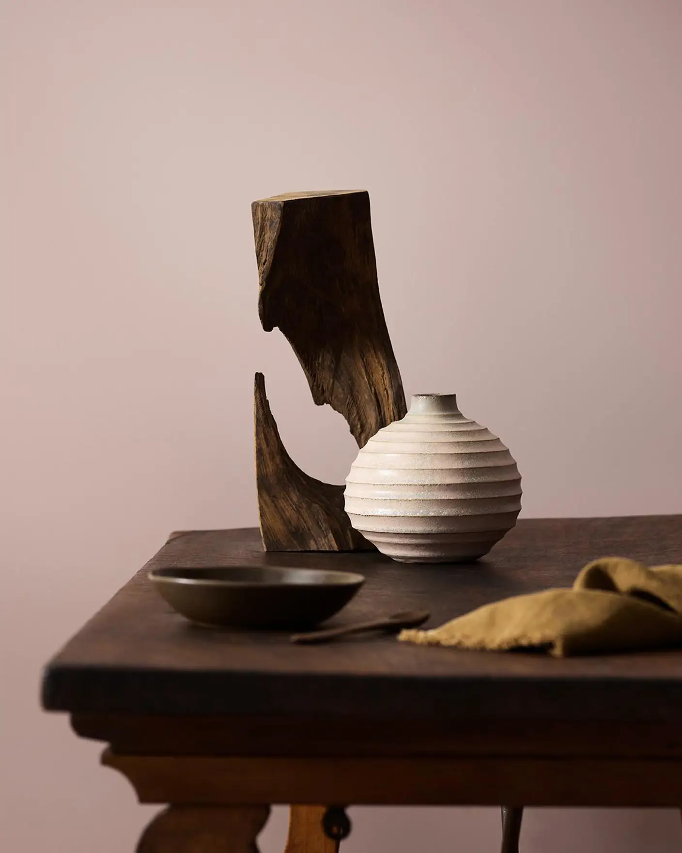 Wooden table with a ceramic vase, sculpted wood, and a dark bowl against a muted pink background painted with the colour 20054 Silky Pink from Jotun.