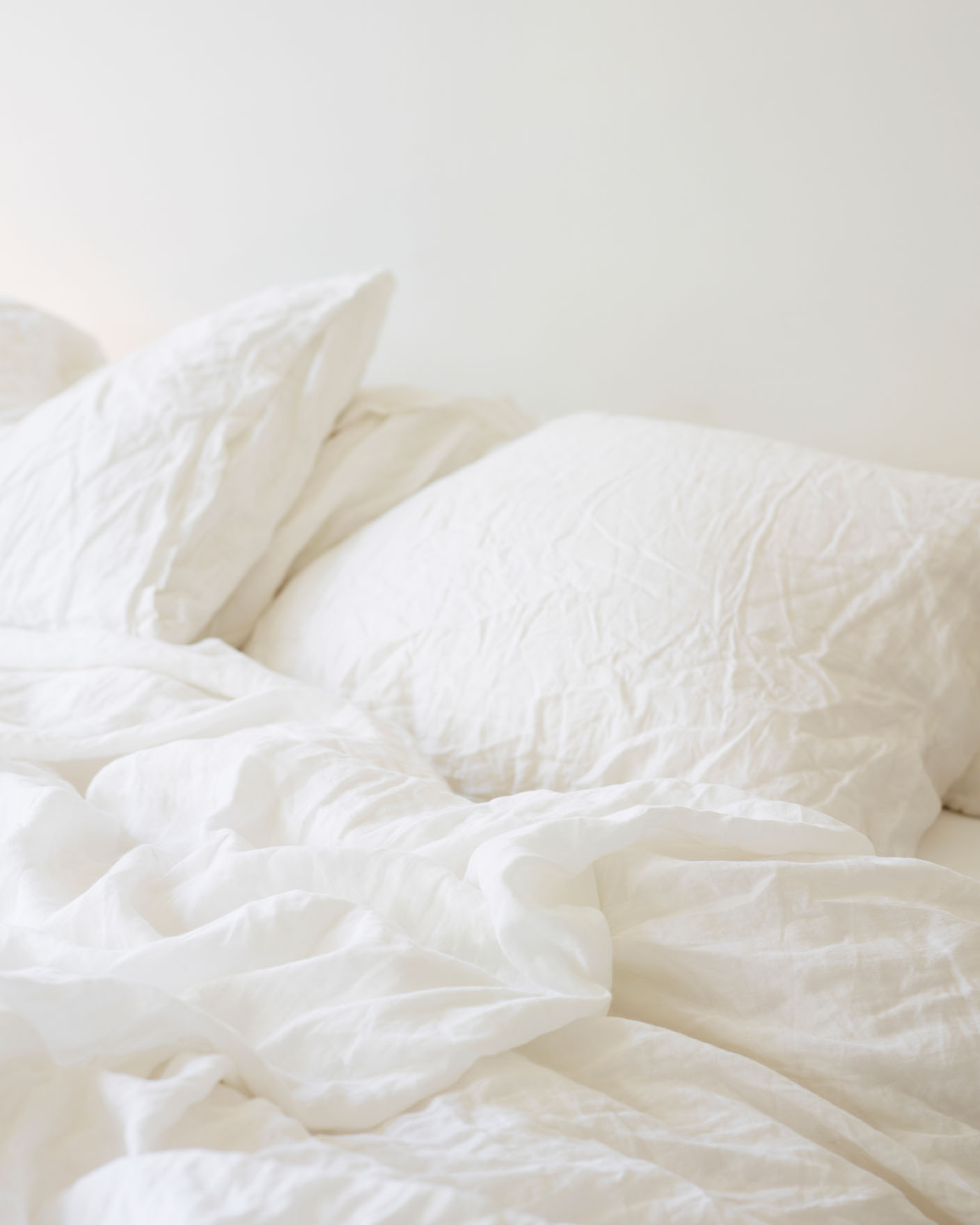 Unmade bed with soft, crinkled white linen bedding and pillows against a warm, grayish wall painted with Jotun 1622 Reflection.