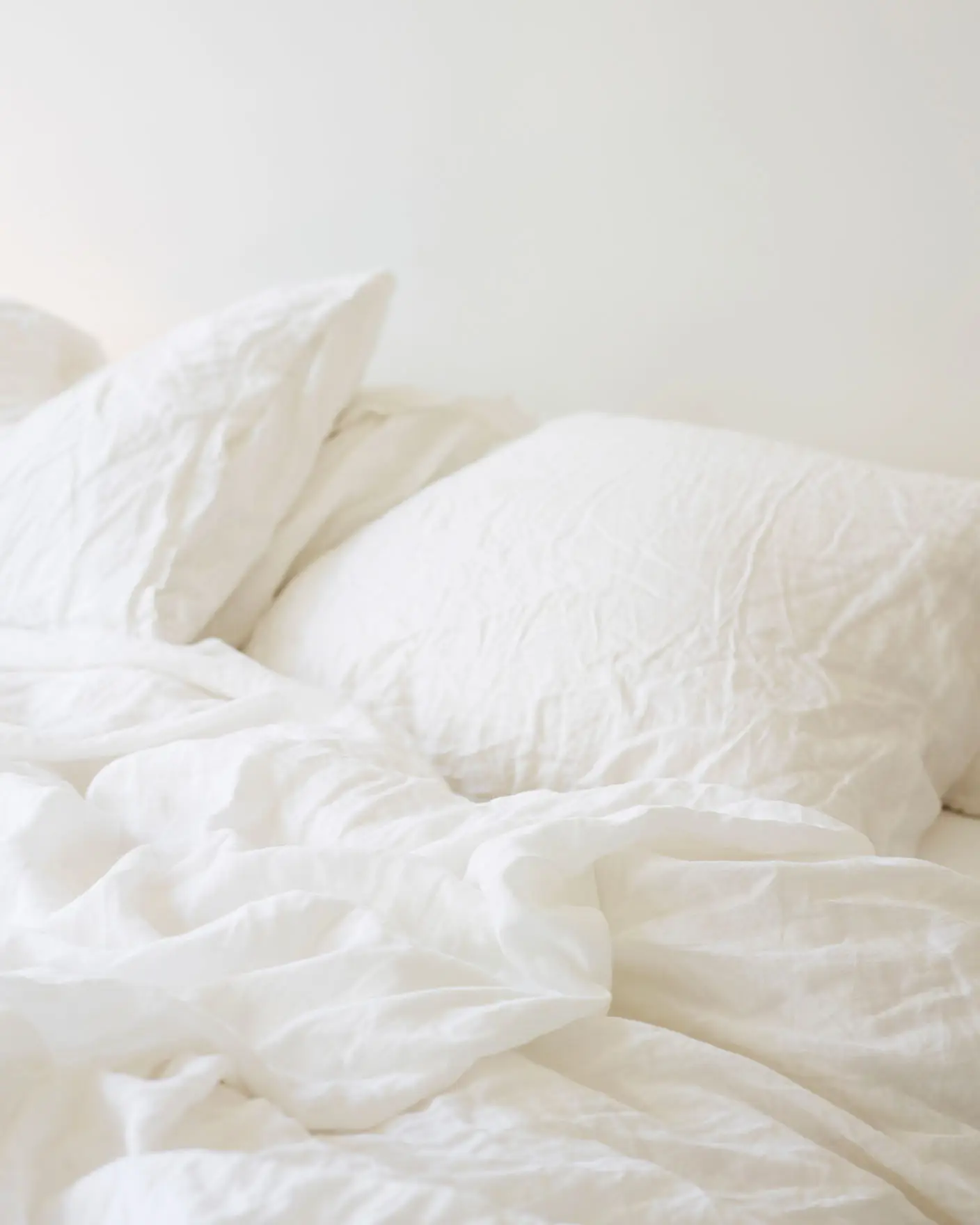 Obäddad säng med mjukt, skrynkligt vitt linnelakan och kuddar mot en varm, vitmålad vägg i Jotun 1622 Reflektion.